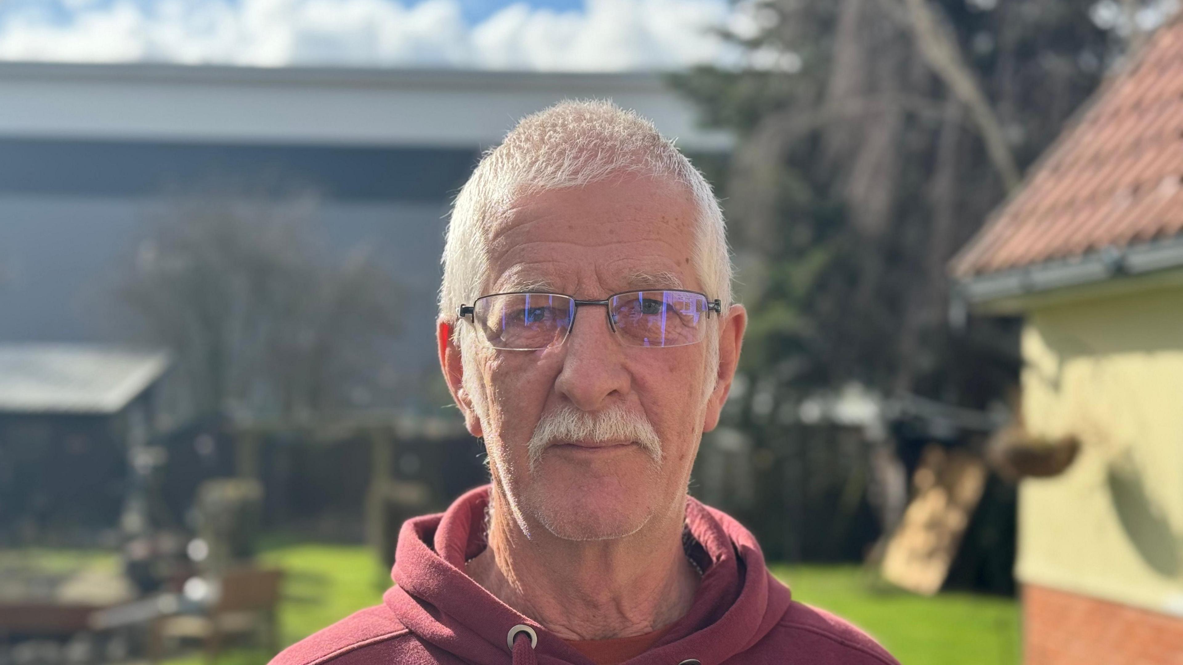 Jim Snell - a man wearing a red hoodie staring into the camera while standing in a back garden. The warehouse can be seen in the background.