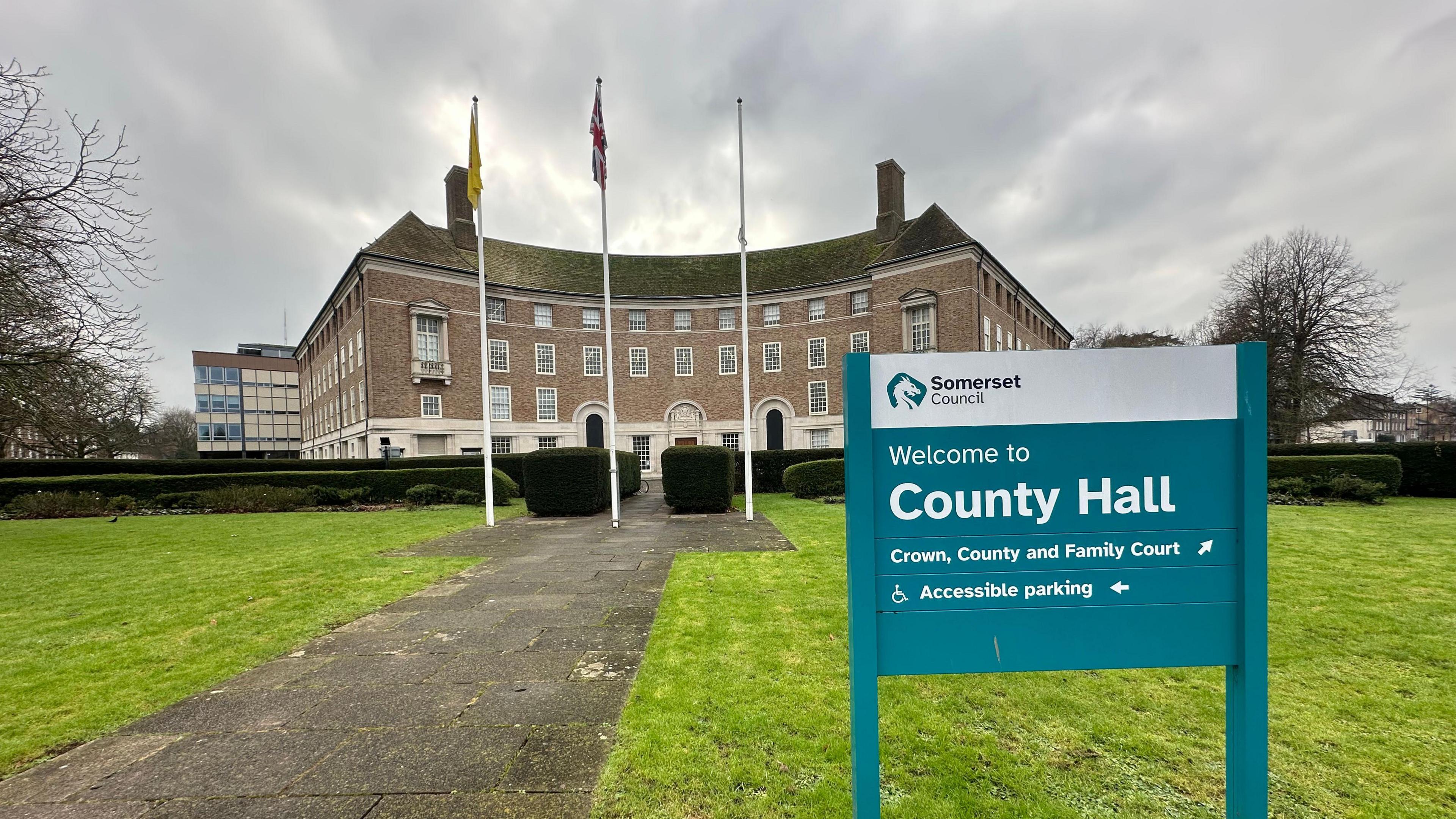 A sign pointing towards County Hall in Taunton, which is in the background. There are three flags, a green lawn and a path in front of the building.