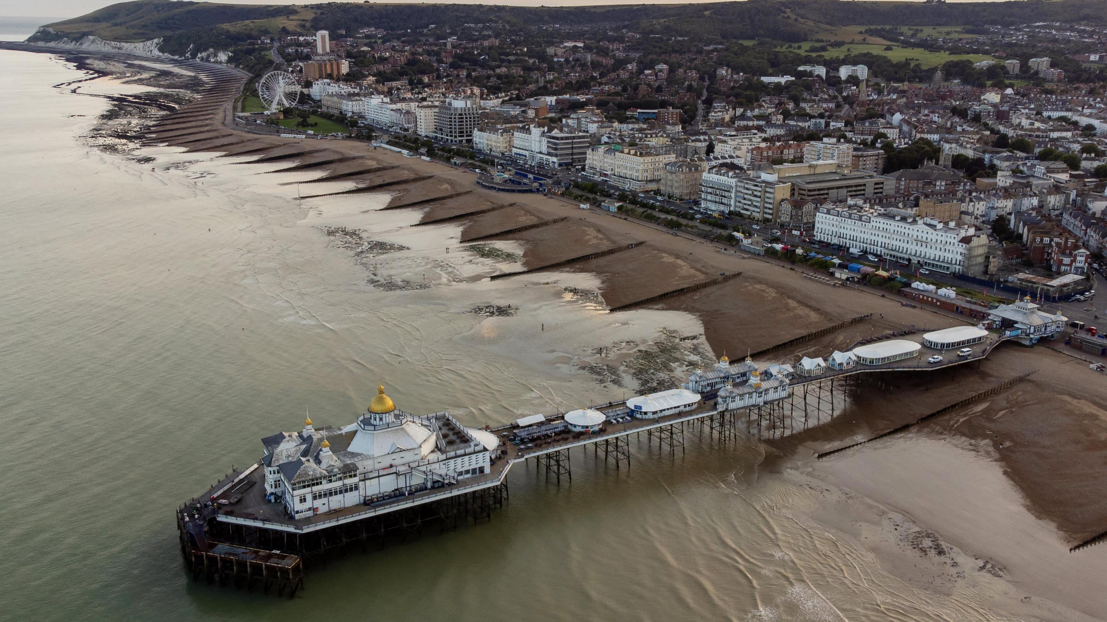 An aerial view over Eastbourne seafront. There is the white pier in the foreground and cliffs and hills in the background.