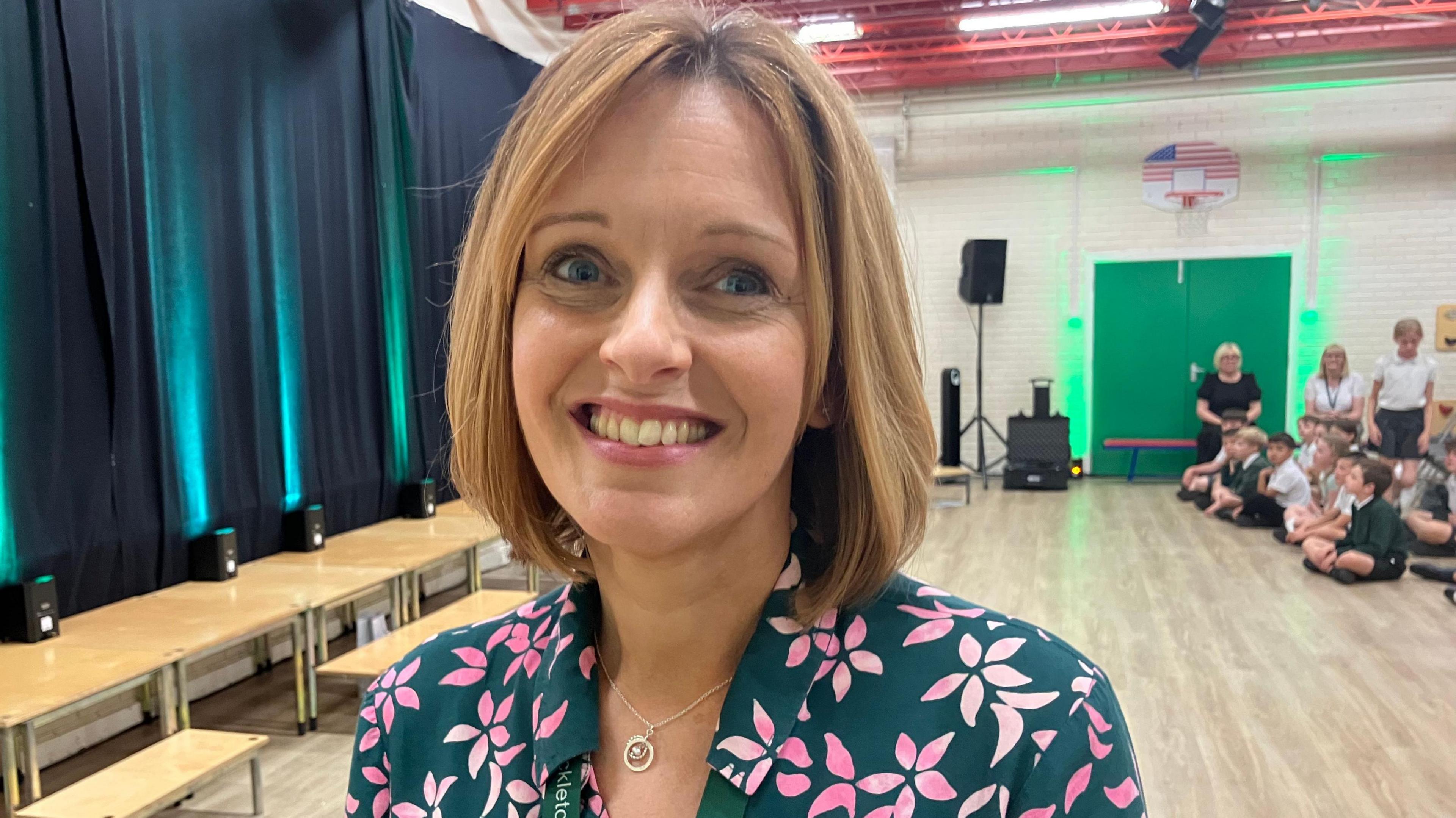 Sarah Gillam, in a green and pink flowery top, wears a lanyard. Her fair hair is cut just above her shoulders. She is standing in a school hall, with children sitting cross-legged in the background.