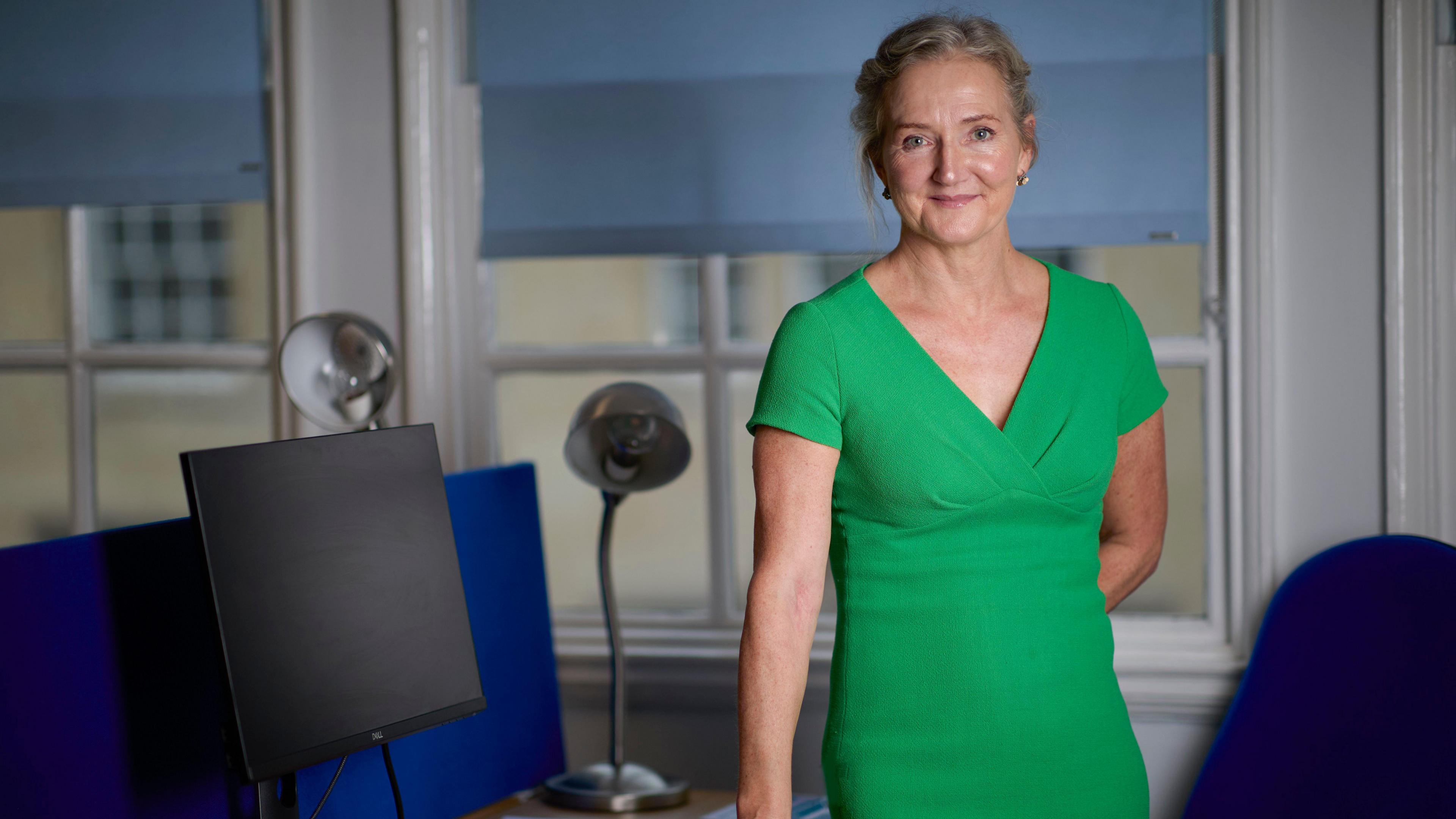 Rosie Phillips is standing at a desk in a green dress. Her hair is up and she is smiling. 