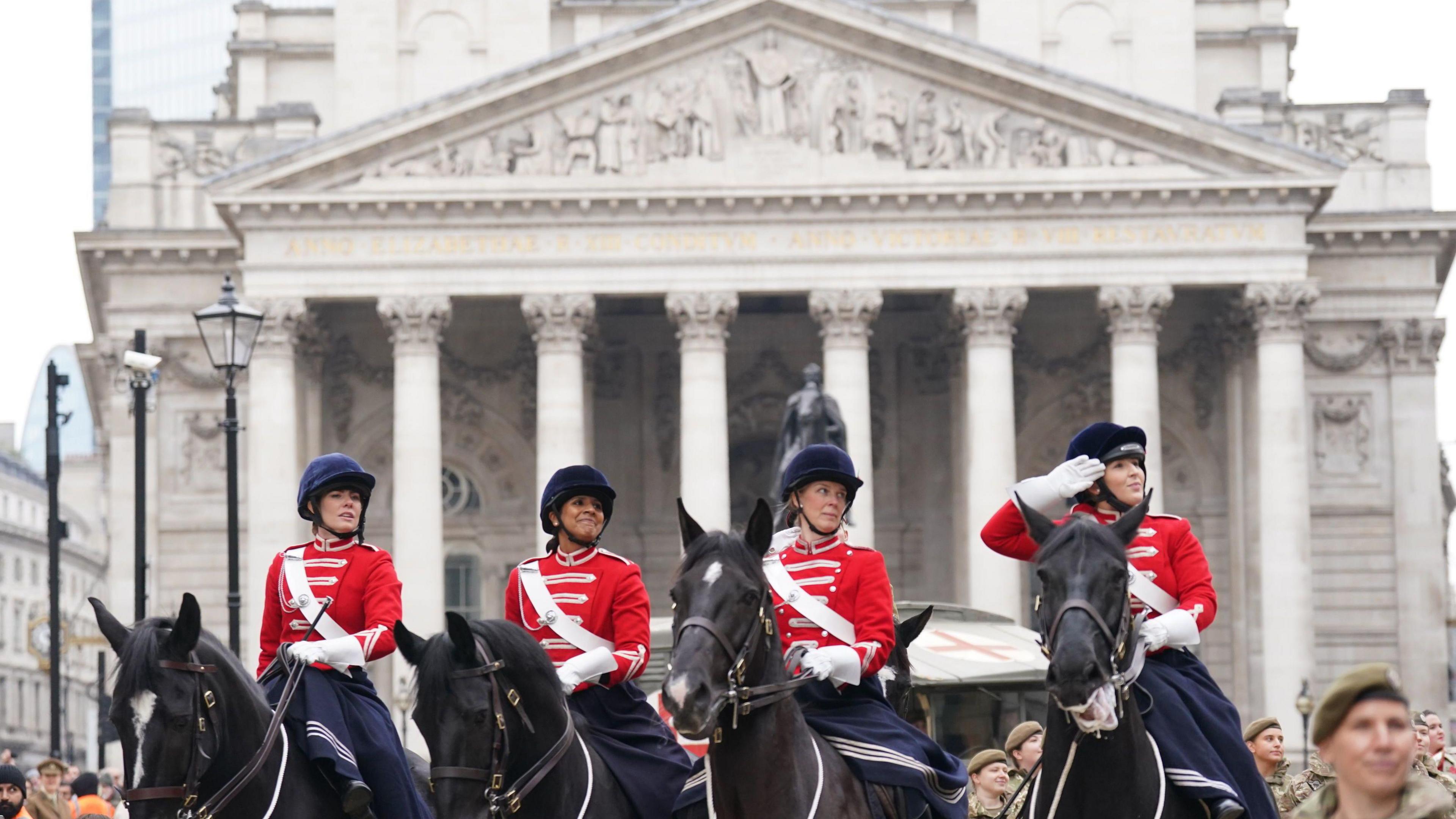 Four women in red military uniforms on horseback participate in the Lord Mayor's Show procession in front of an ornate classical building, with columns and intricate carvings. One rider is saluting, and the group is surrounded by spectators and other uniformed personnel.