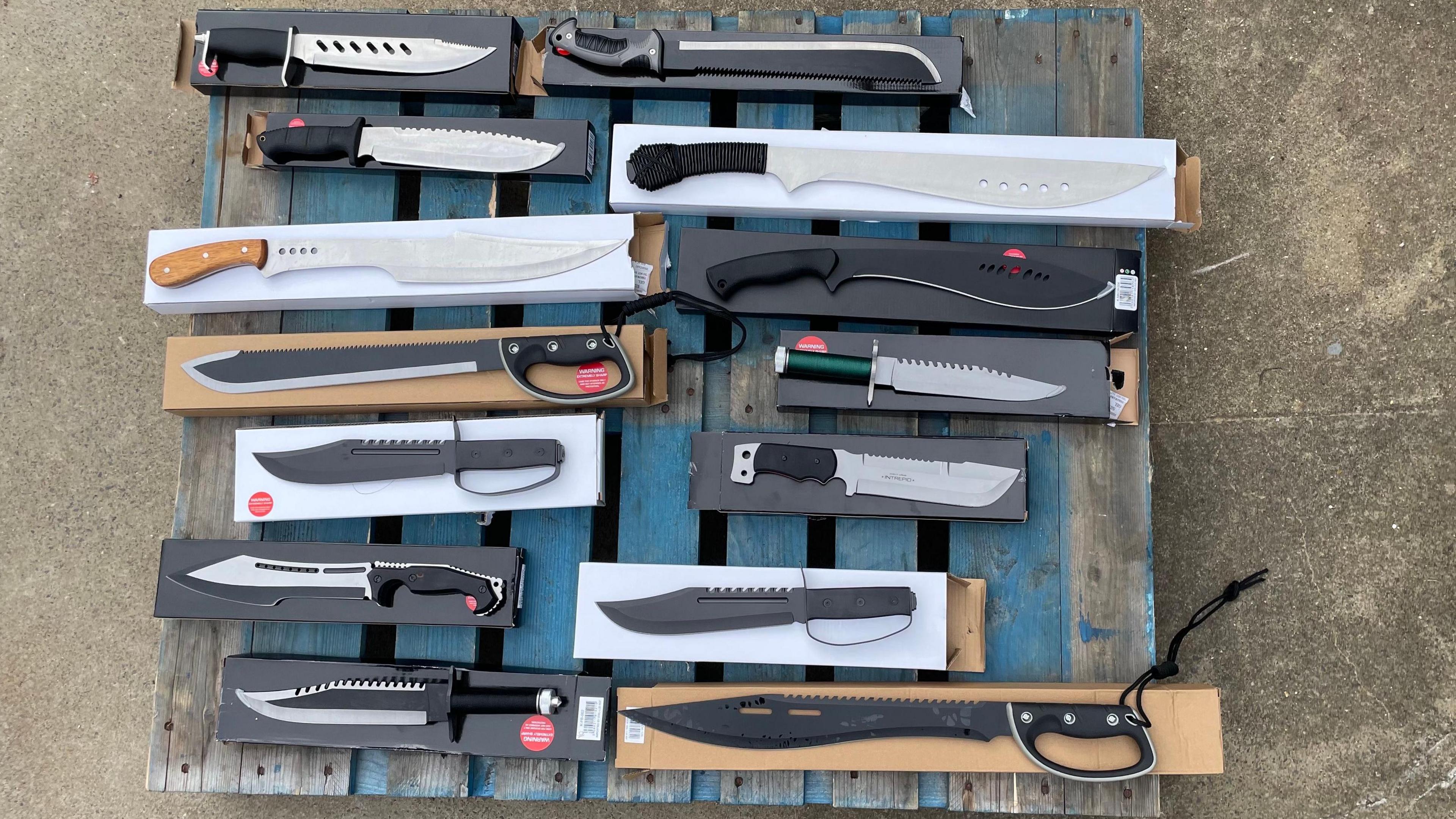 A collection of 14 models of knife lying on top of their individual boxes, which are in turn laid out on a blue wooden pallet. The knives are all flat and are pictured from above. All the knives meet the criteria of the new ban, having blades longer than 8in or 20cm, as well as other features like holes in the blade or serrations.