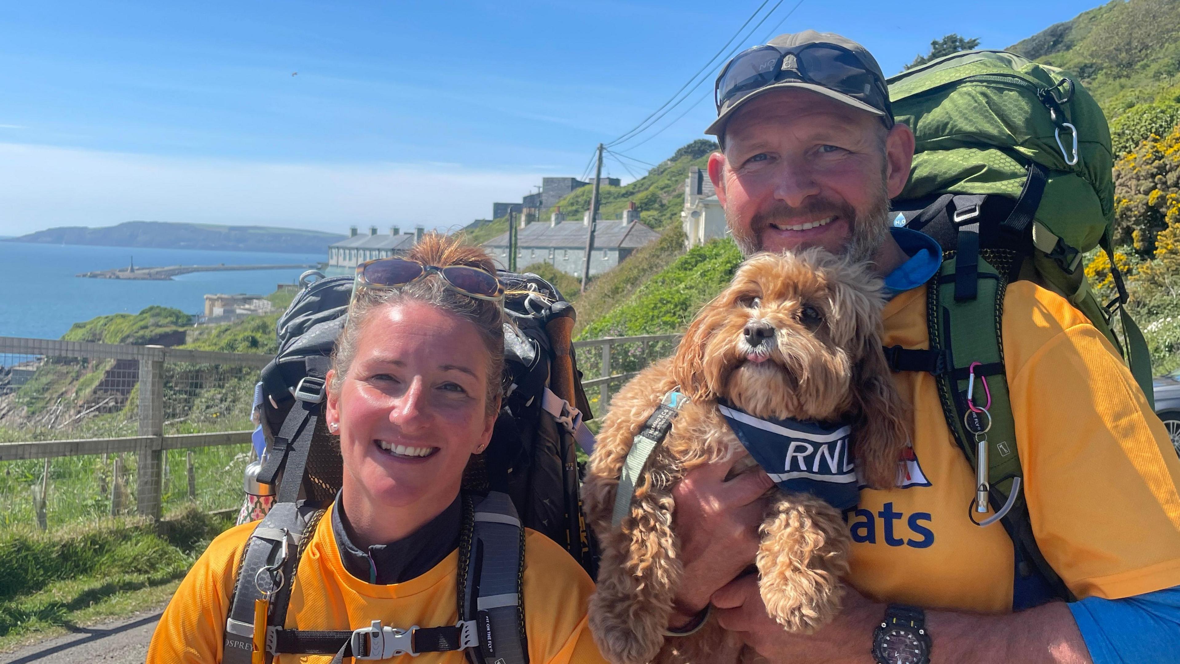 A couple in bright orange t-shirts that say RNLI lifeboats smile at the camera. Jason is holding their dog Teddy who is also wearing an RNLI bandana around his neck. The couple have huge backpacks on their backs and the coast path is behind them.