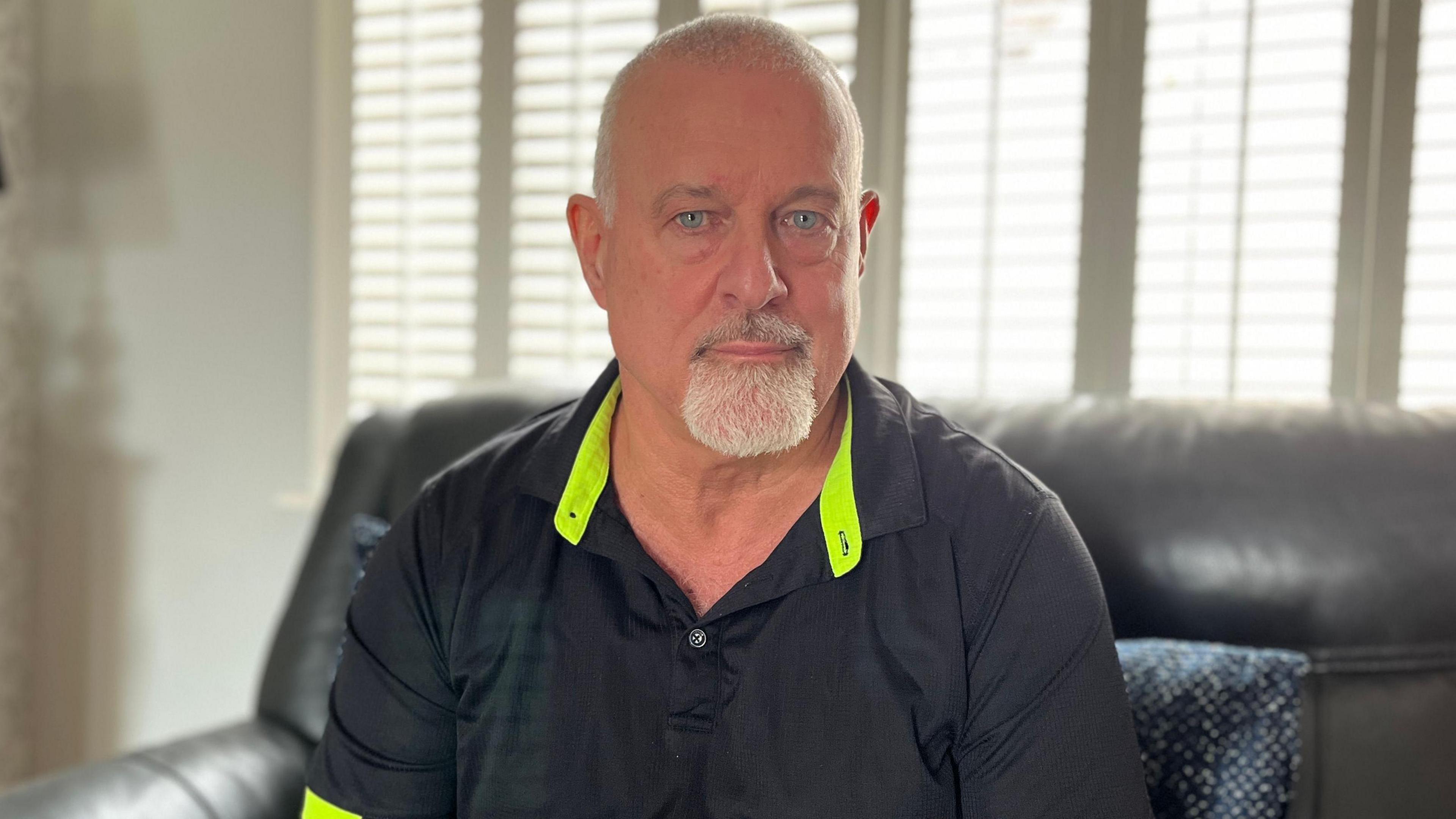 Craig Nairn sits in his living room, wearing a black shirt with neon collar