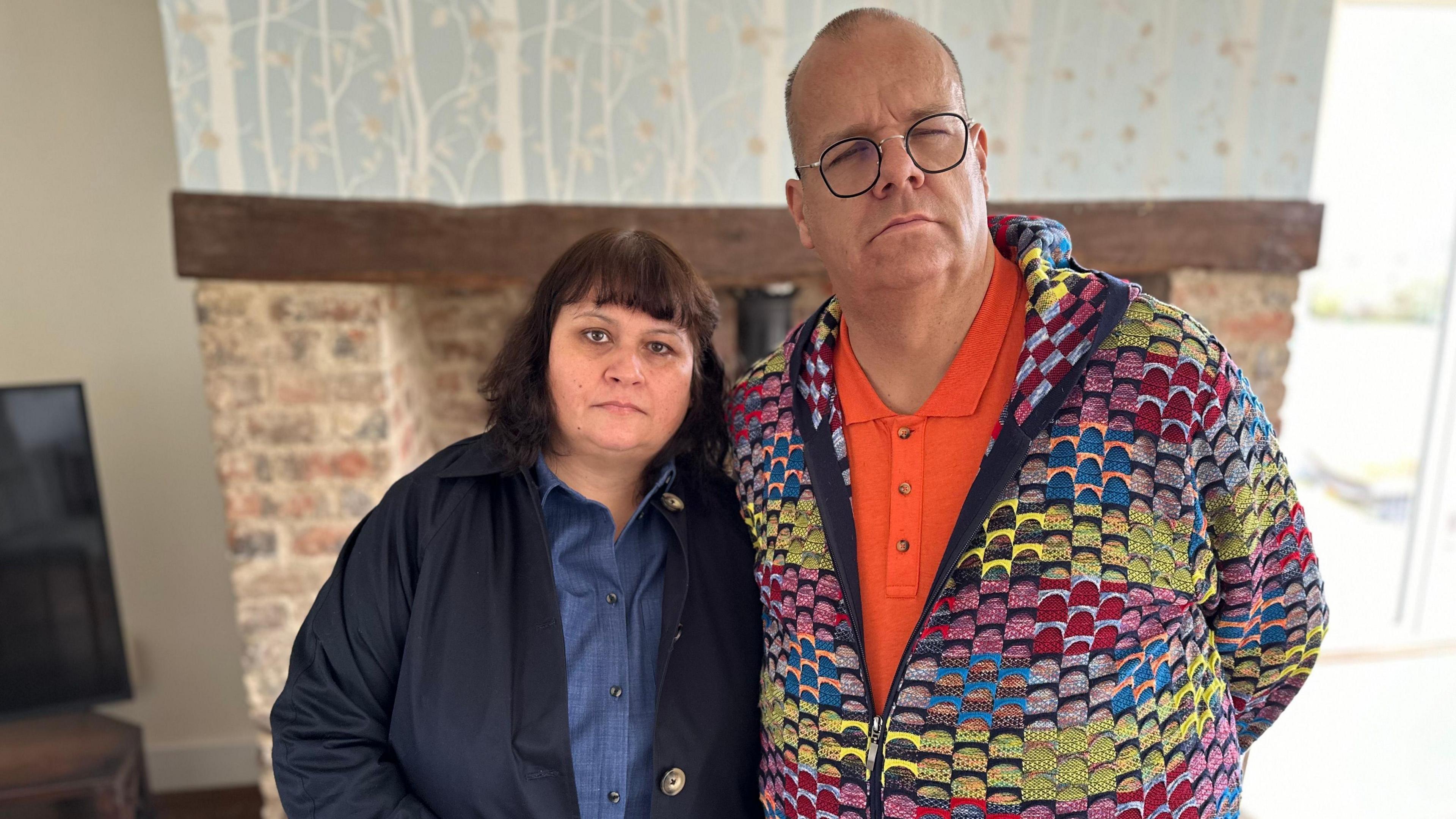 Gary and Susan Dowers arm in arm. Susan has dark hair in a bob with a fringe. She wears a navy shirt and jacket. Gary has closely cropped hair and glasses. He wears an orange t shirt with a colourful cardigan.