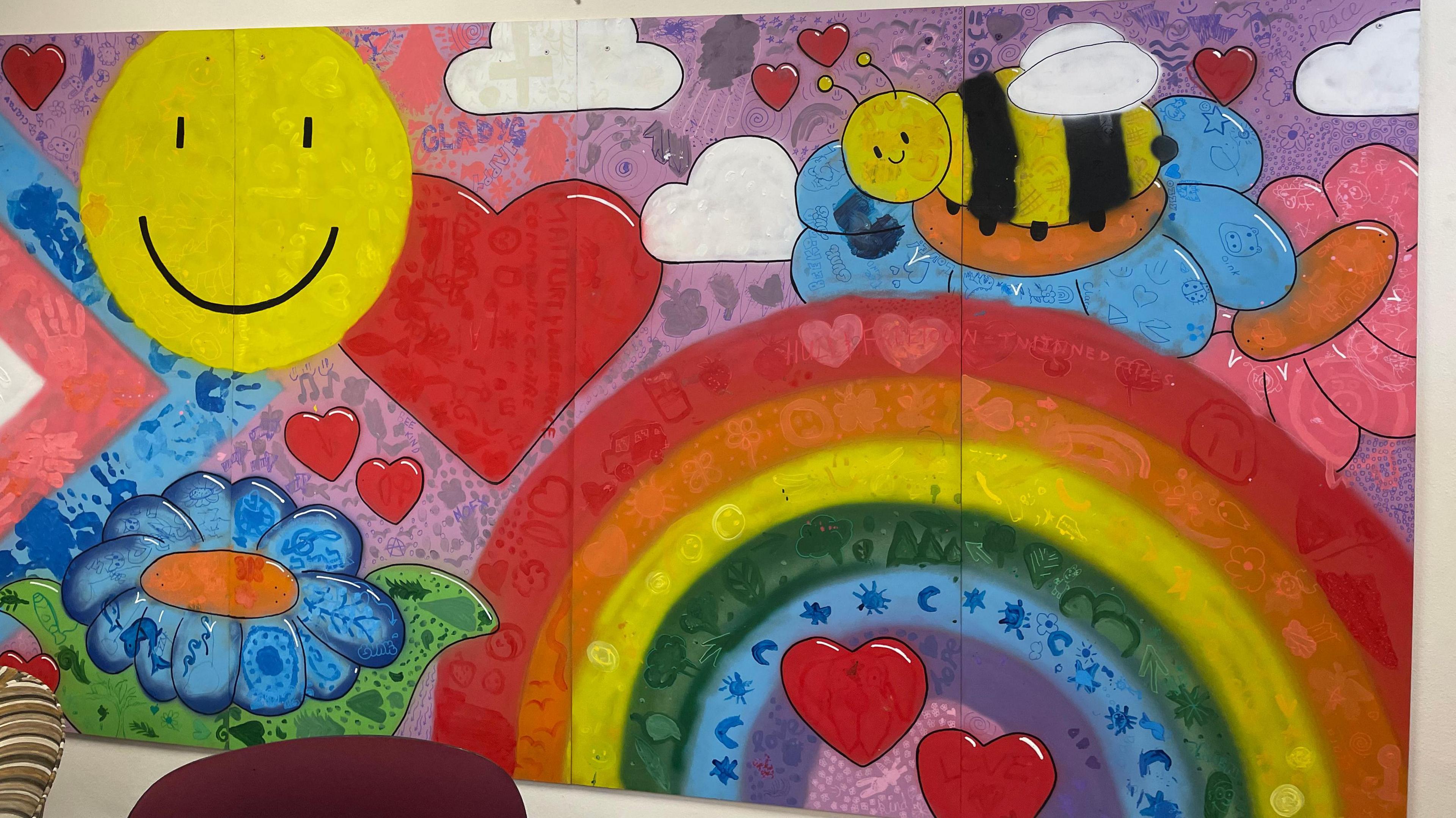 A colourful painted mural includes red love hearts, a big yellow smiley face, clouds, a rainbow, flowers and a black and yellow bee, along with a series of handprints.