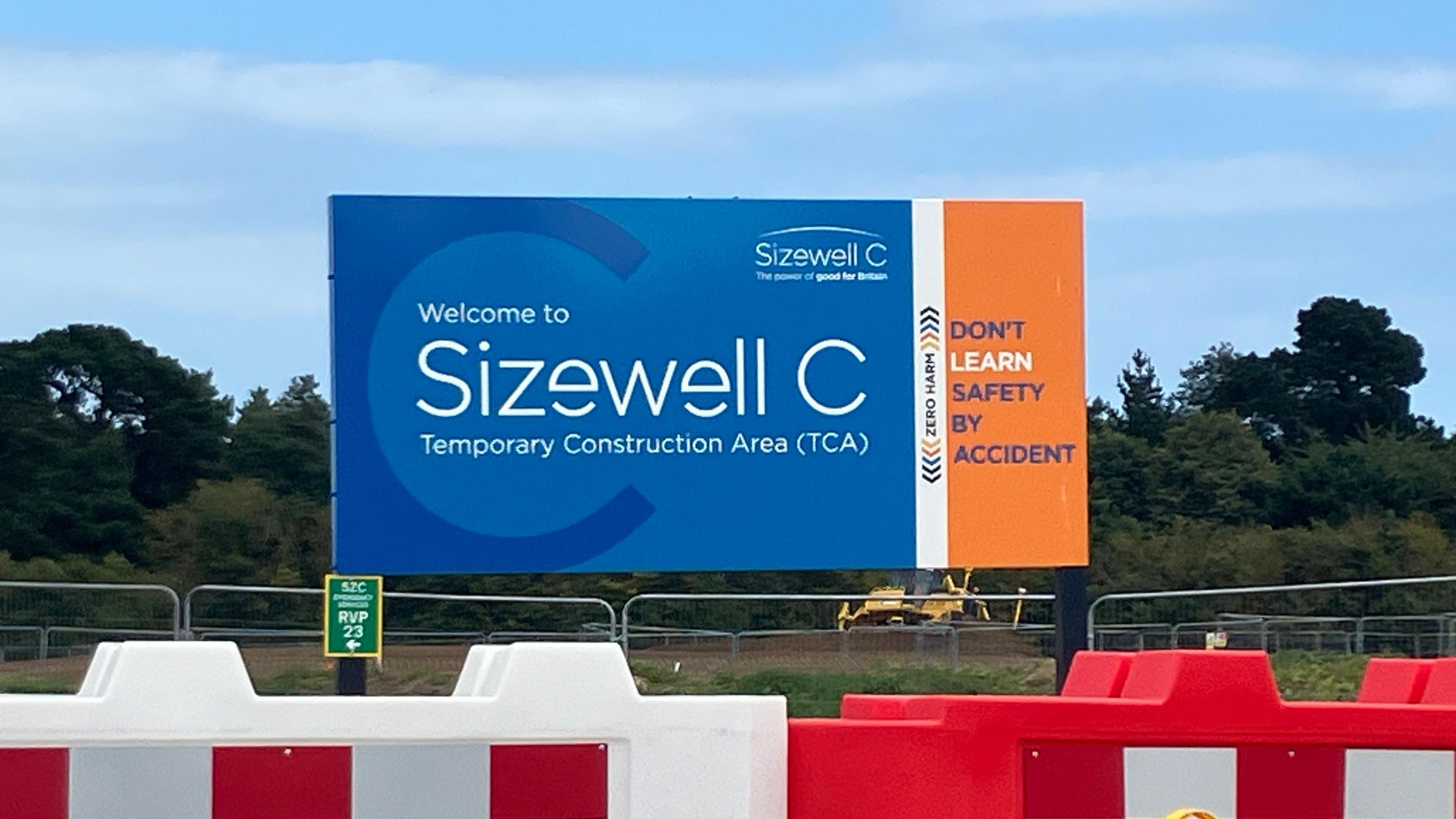 A Sizewell C board stands alongside a road. Red and white fences sit in front of it.