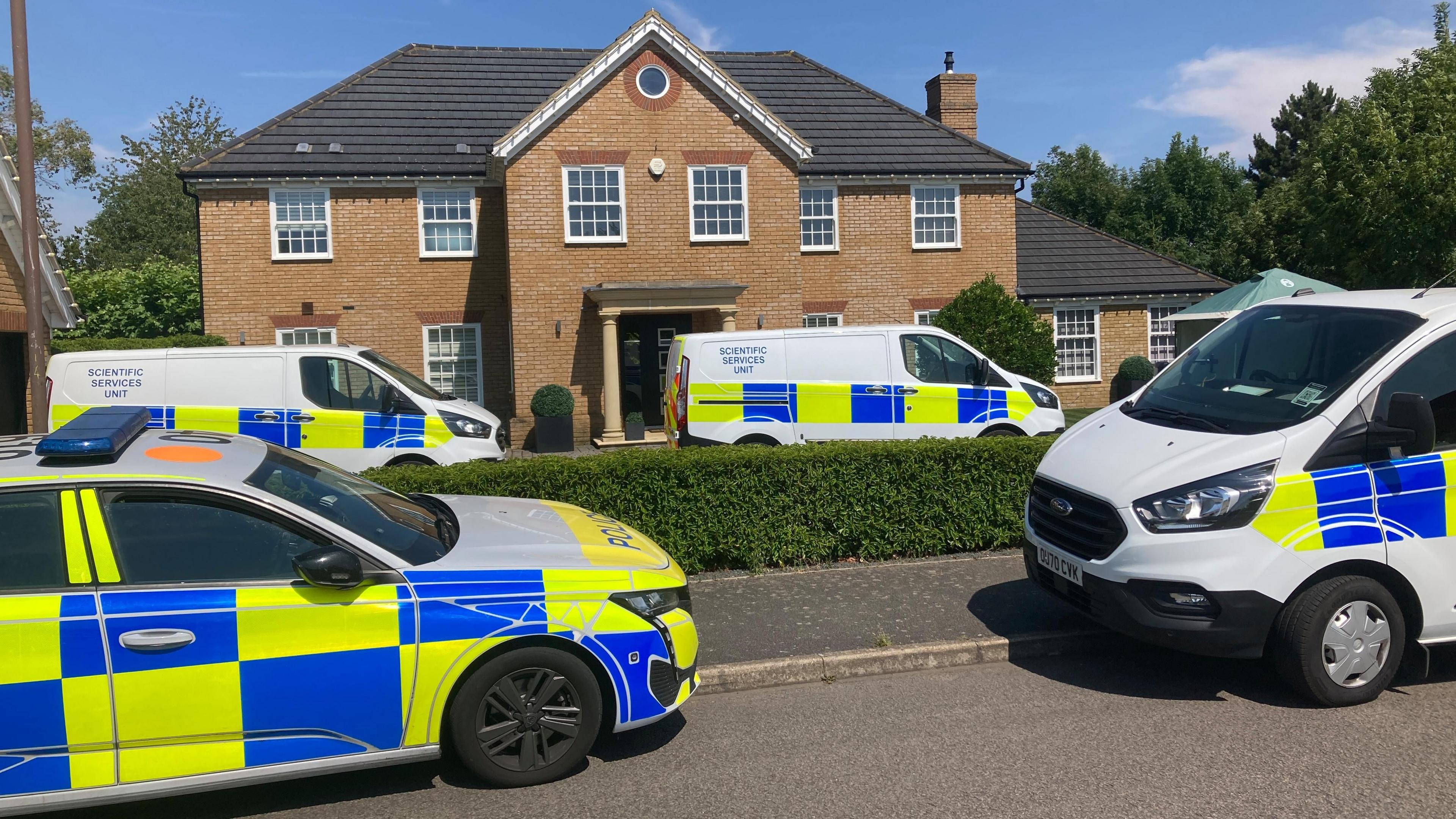A police car and a few forensic vans are parked outside a large house – some on the street, some on the driveway.