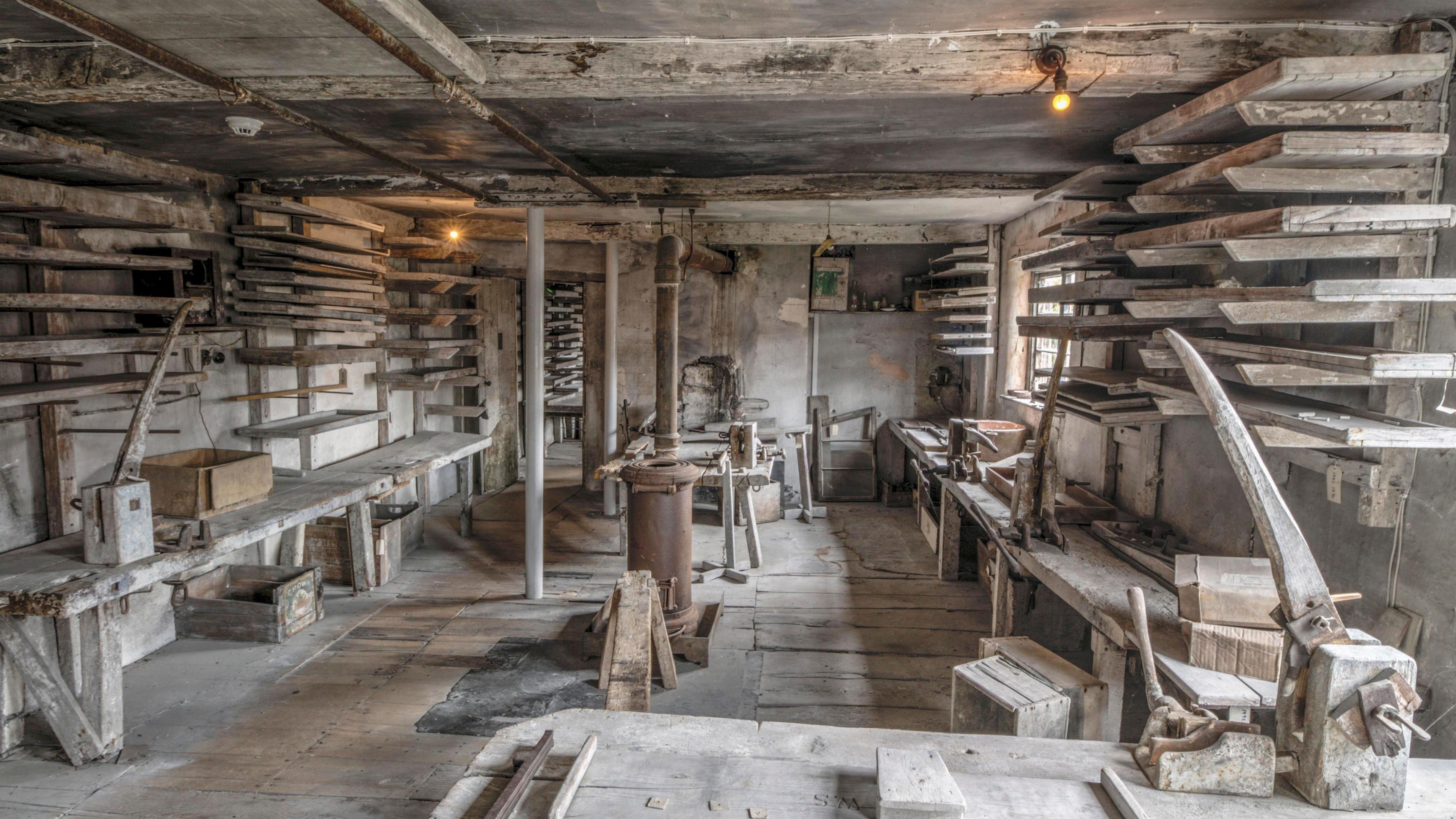 The interior of the Pipeworks. A fairly small wooden room, with large beams on the ceiling and workbenches along both sides of the room. In the foreground there is a bench with what looks like a kind of vice on it. The sides of the room have rows and rows of shelves. Everything is bathed in a grey-beige dust from the clay. There is light coming in through a window towards the back of the room. 