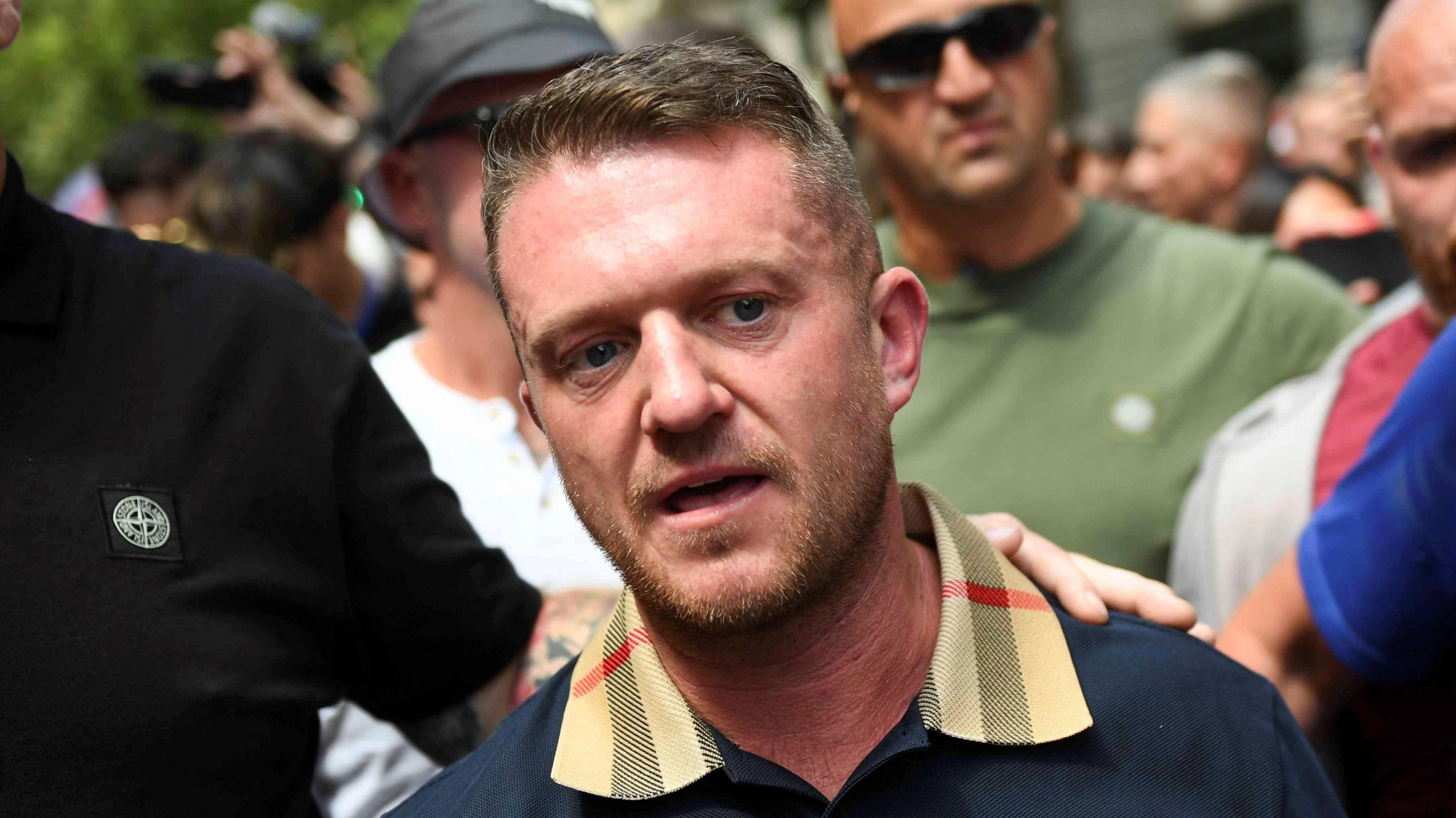 Tommy Robinson at a protest in July. He is in a crowd of people wearing a navy top with beige collar.