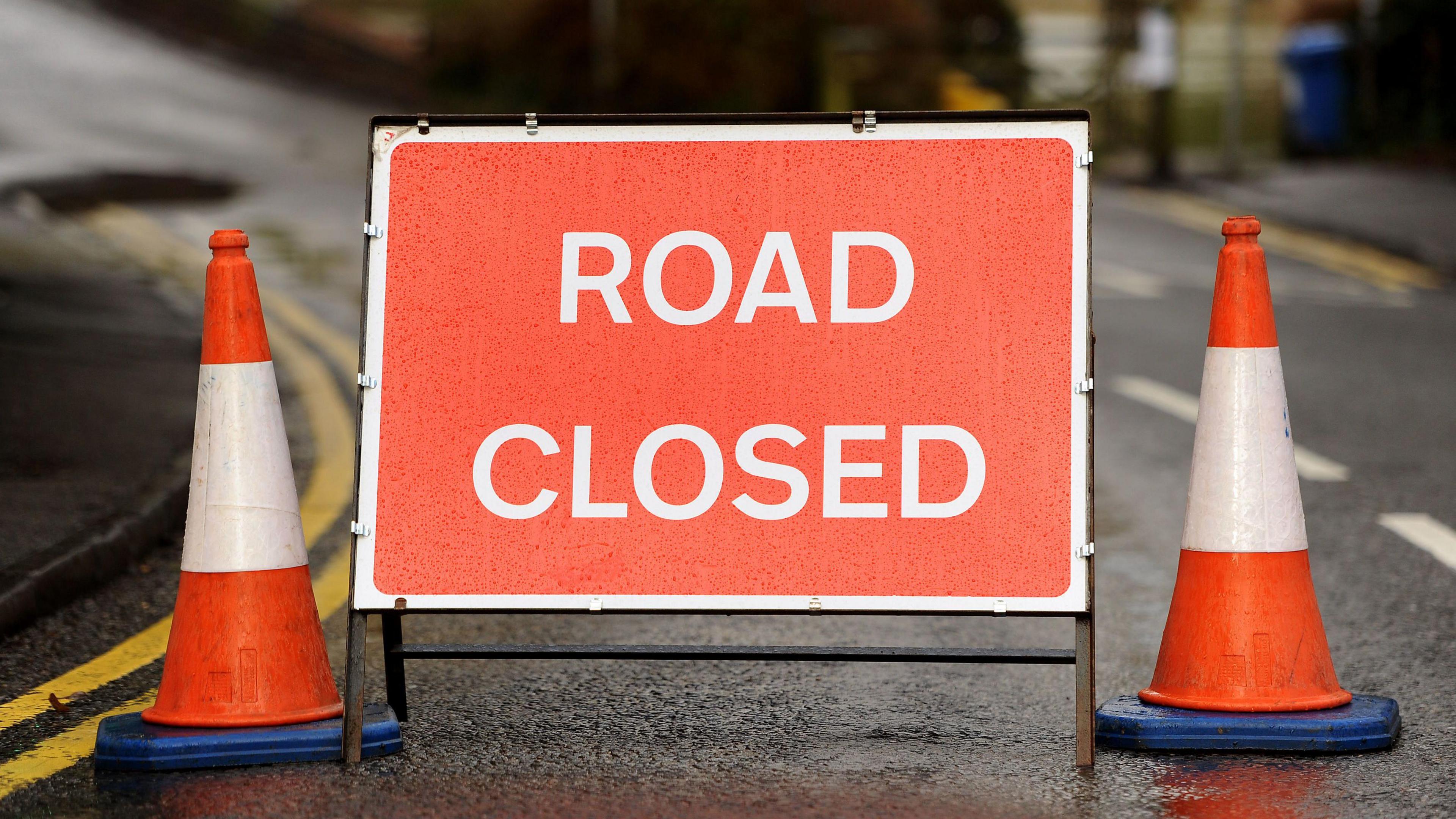 A red-orange "road closed" sign, between two traffic cones