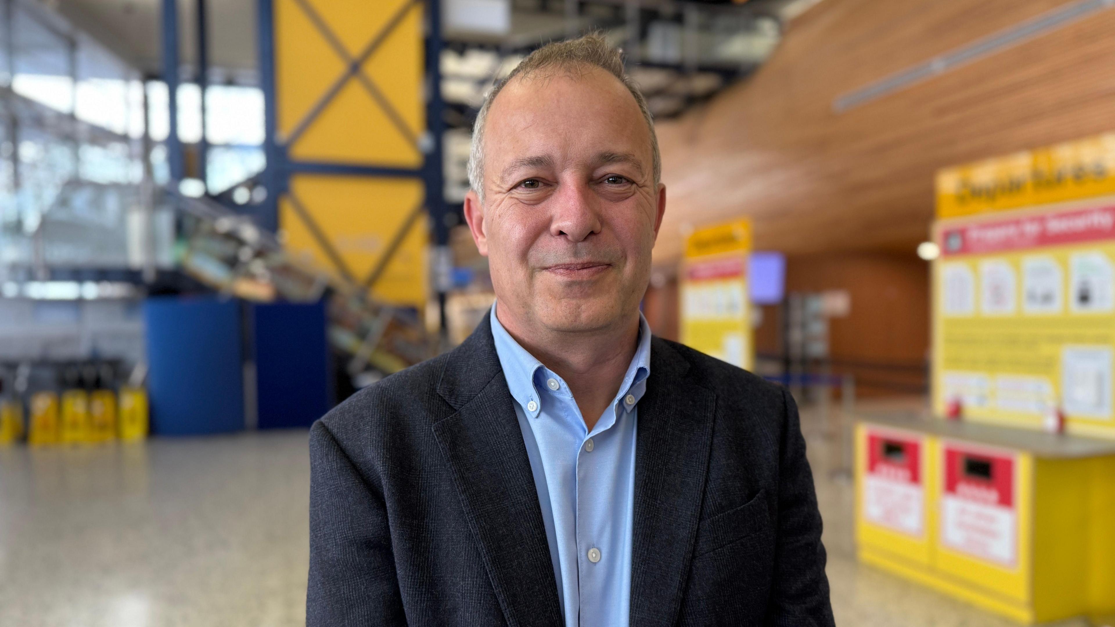A man smiling inside Guernsey Airport, wearing a suit and shirt.
