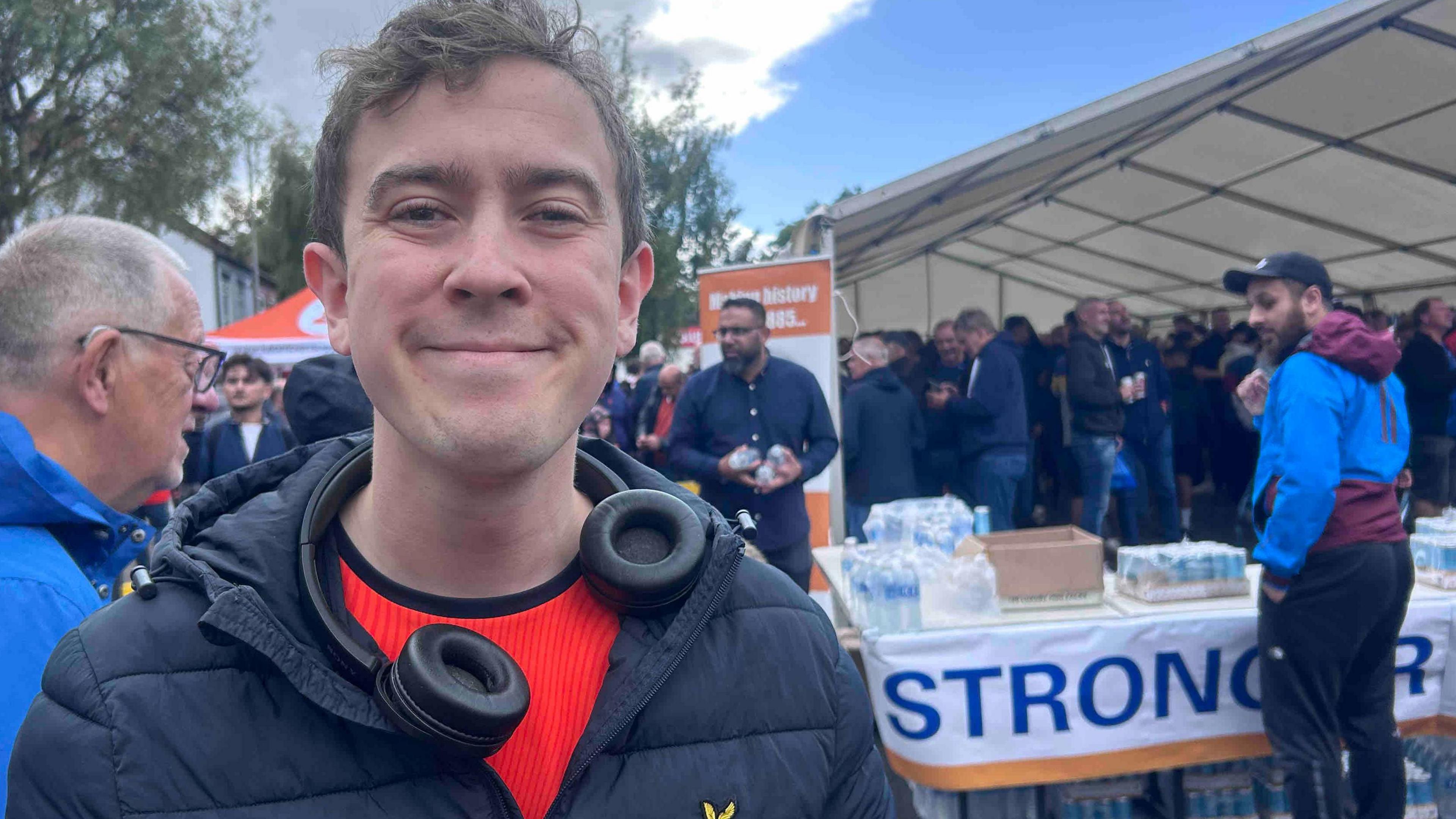 Alex Teunion has light brown hair and is wearing an orange T-shirt and black puffa jacket. He is smiling at the camera and has a pair of headphones around his neck.