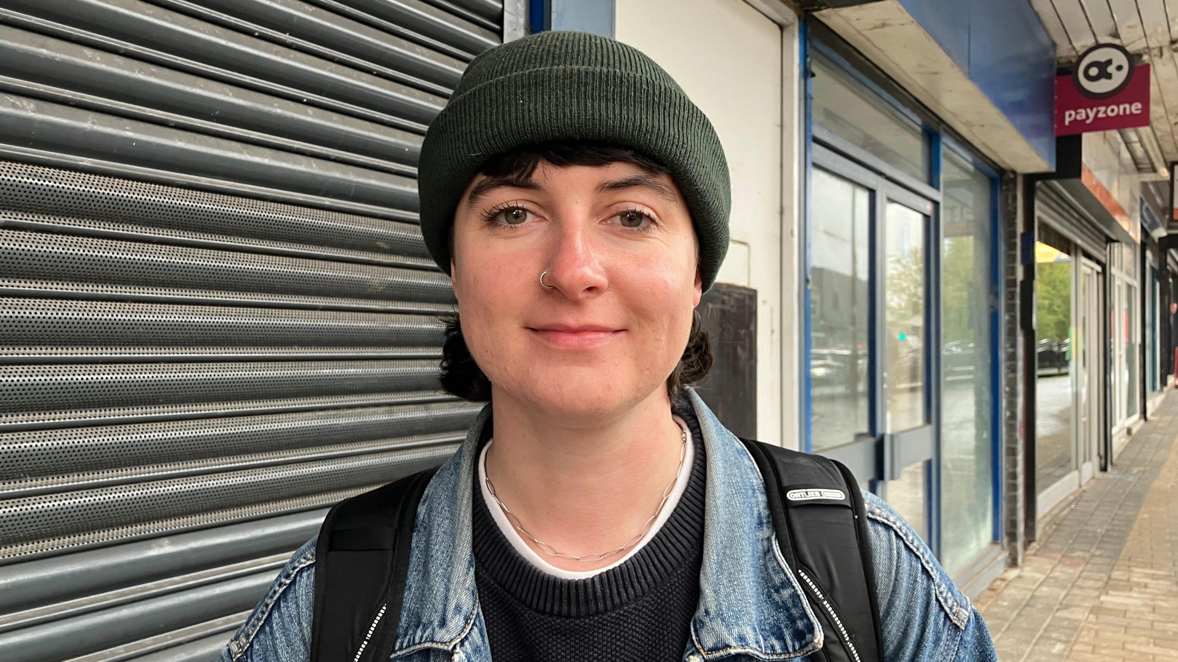 A person smiling at the camera, wearing a dark green beanie hat, black jumper and a denim jacket. In the background are shut down shops.