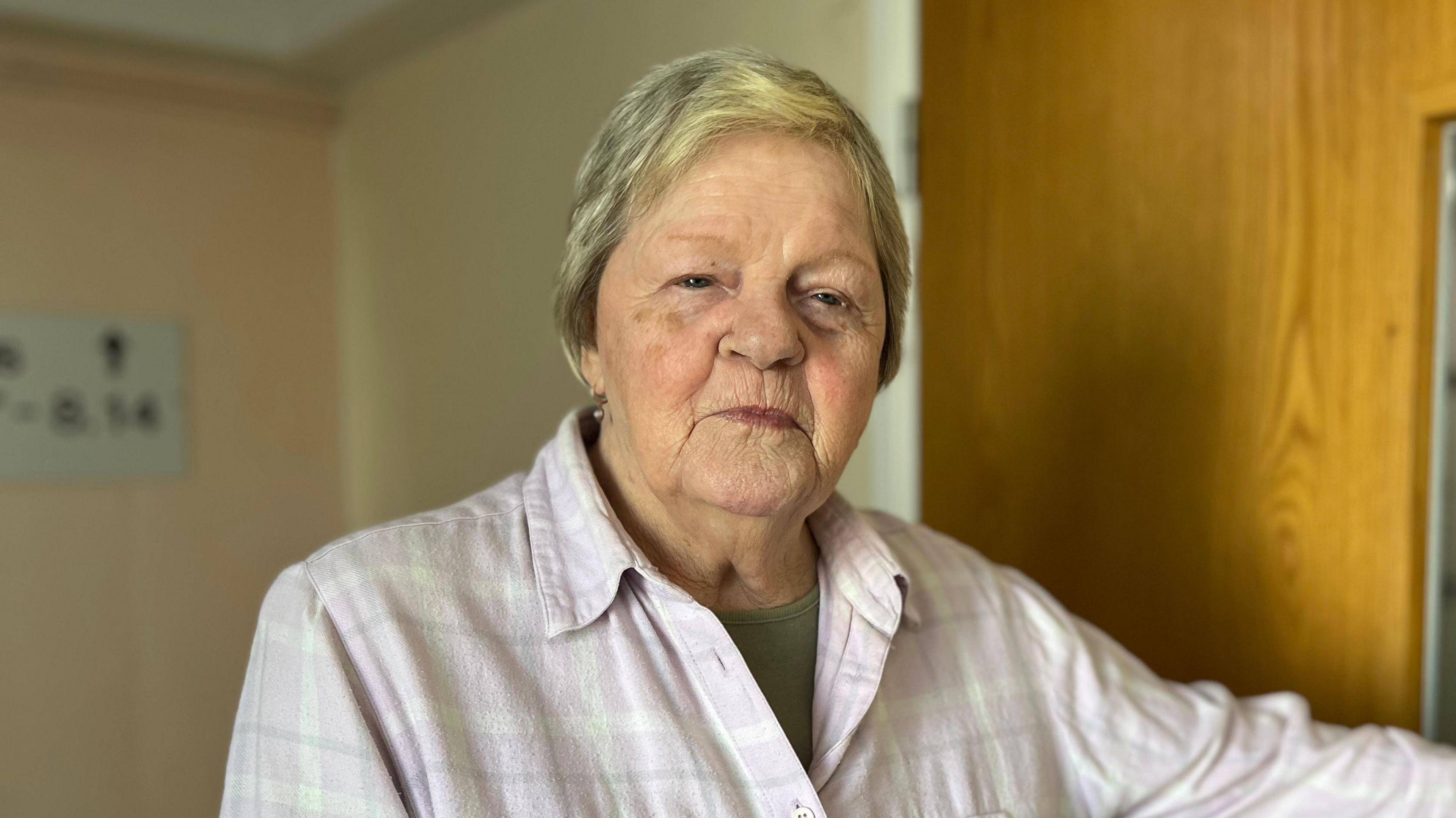Julia McBride, an 81-year-old white woman with cropped blonde, greying hair wearing a light pink flannel shirt revealing a khaki t-shirt underneath.