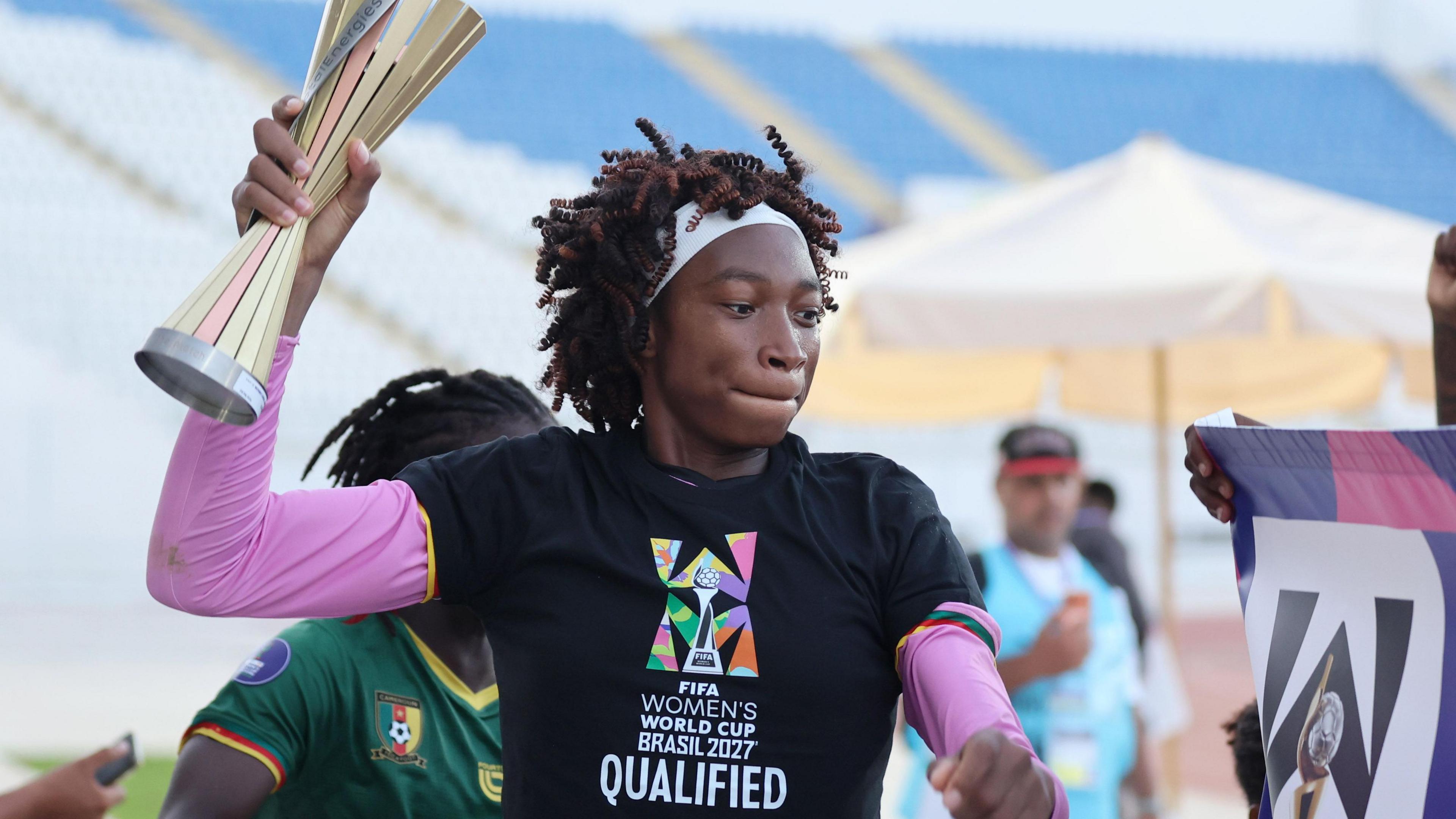 Cameroon goalkeeper Michaely Bihina, visible from stomach up, wears a black t-shirt bearing the message 'Fifa Women's World Cup Brasil 2027 qualified' over a long-sleeved pink top. She has a thick white headband holding back her hair as she holds up a player of the match trophy, which is disappearing out of the top of the image, with her right hand. A team-mate in a green Cameroon jersey is partially obscured behind her, while blurred in the background a man in a bright blue tabard watches on in front of the empty stand of a stadium