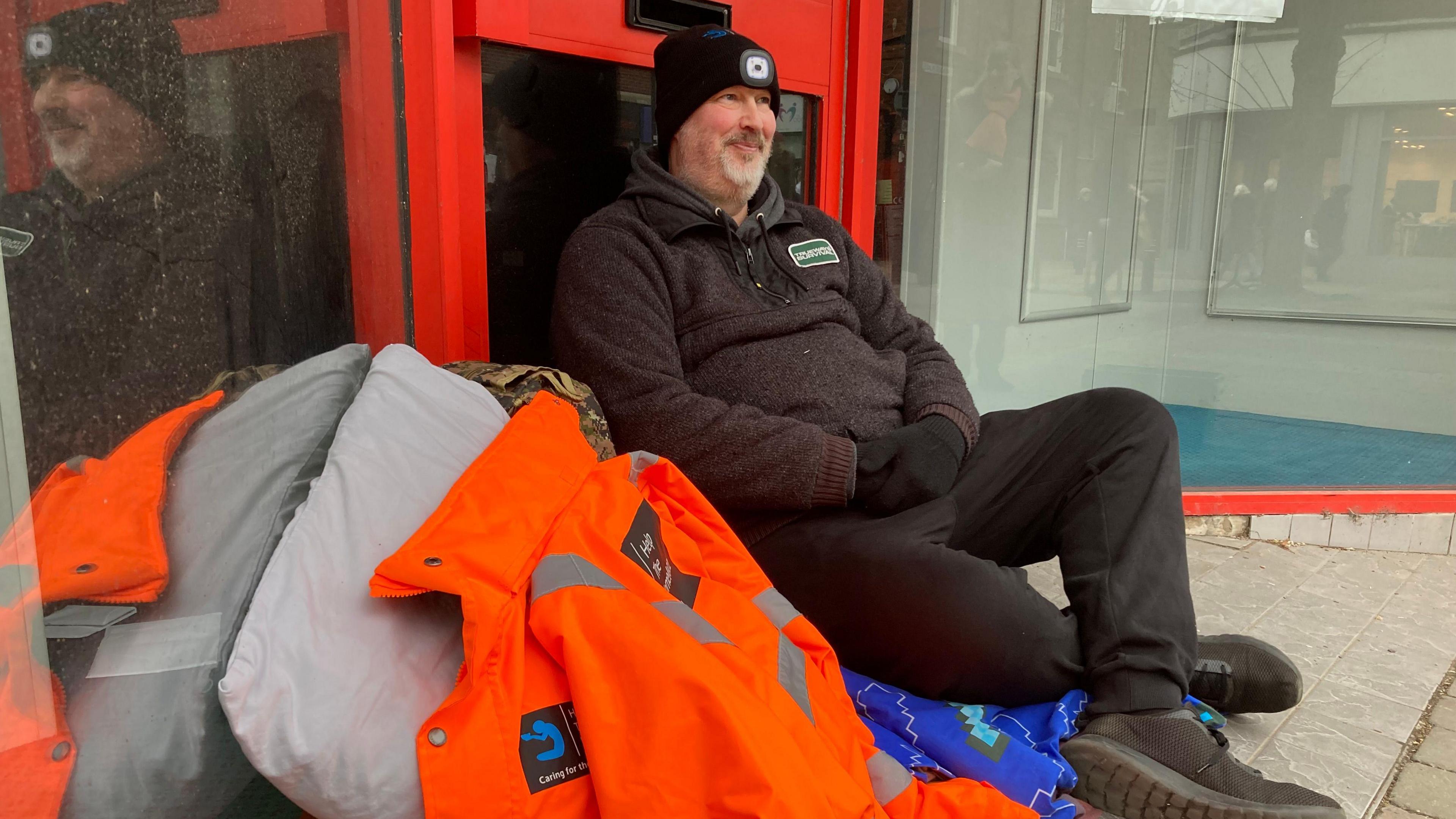 A middle-aged man dressed in black clothes, a black woollen hat and black gloves sits on a blanket on the ground outside a red shop doorway. To his right are an orange jacked, a grey sleeping back and a grey pillow.