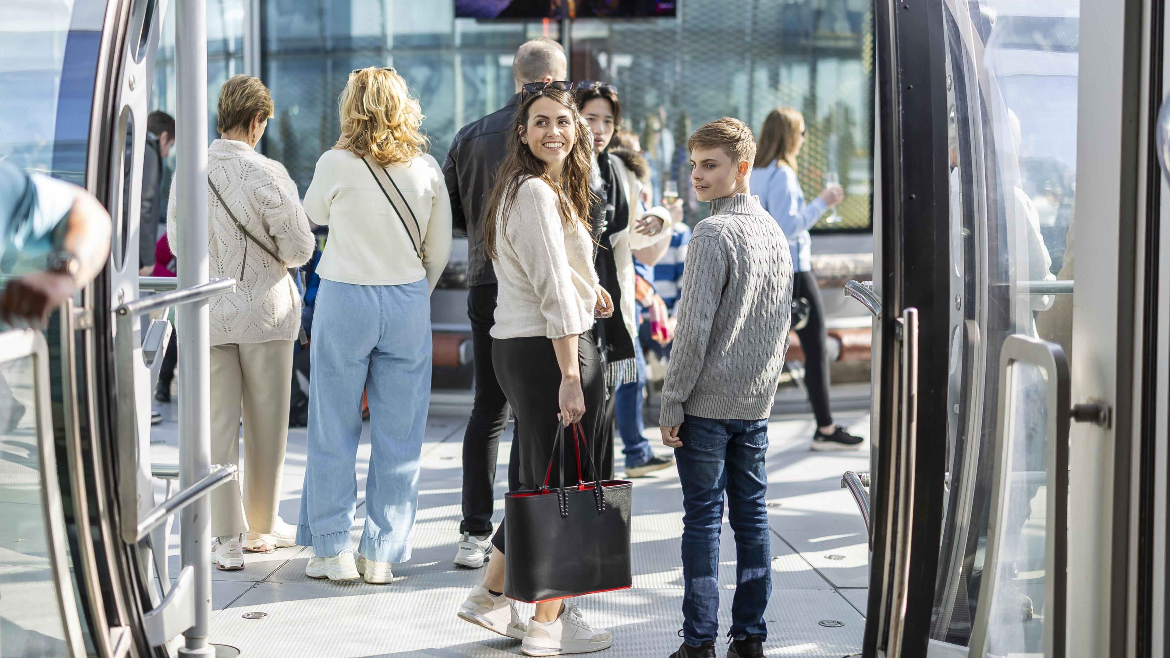 A crowd of people enter a pod with windows and metallic railings. A brunette girl looks over her shoulder and smiles in the entrance.