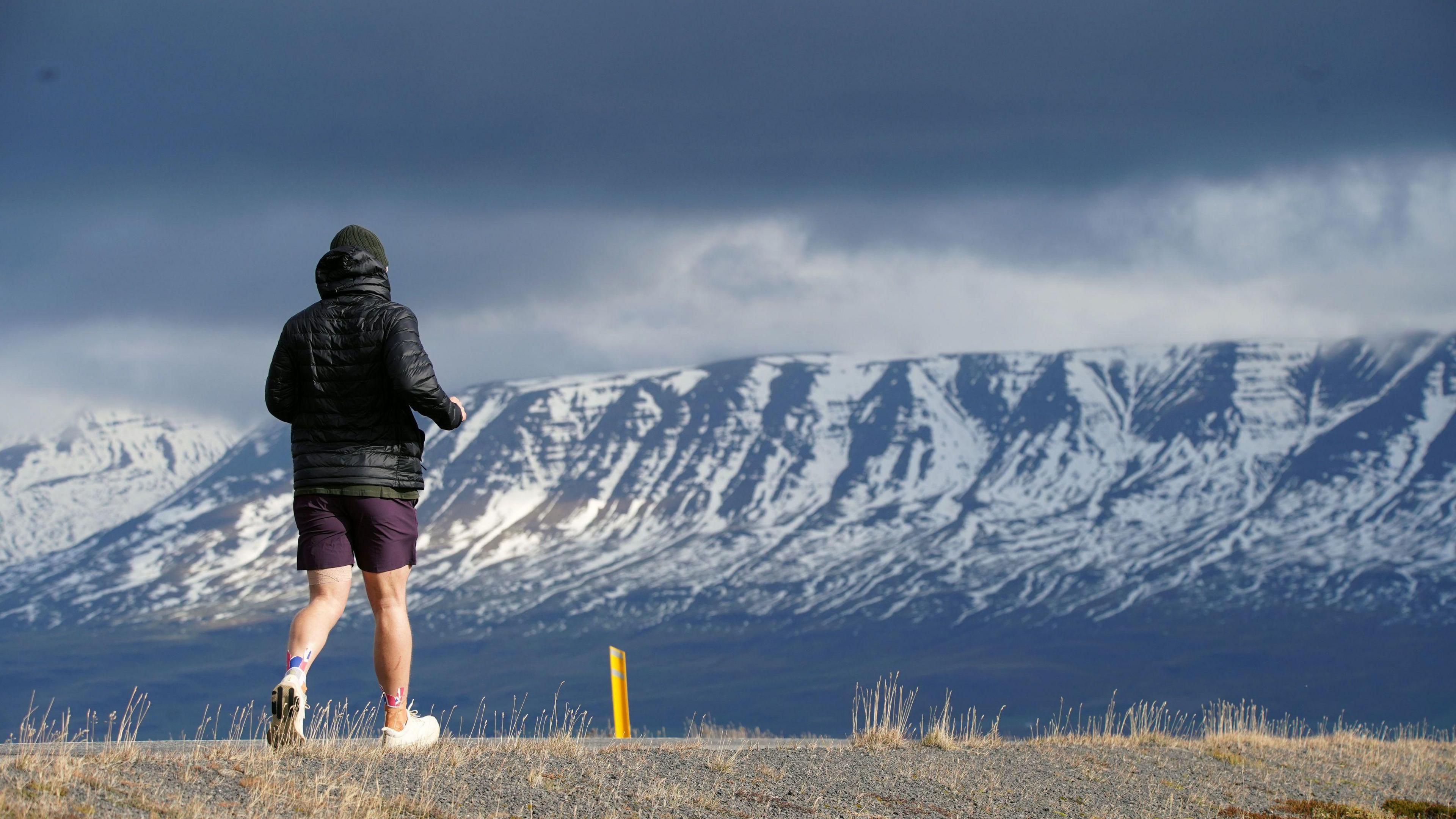 Seb Key running with his back to the camera. In front of him is an epic looking mountain range, with snow on it.