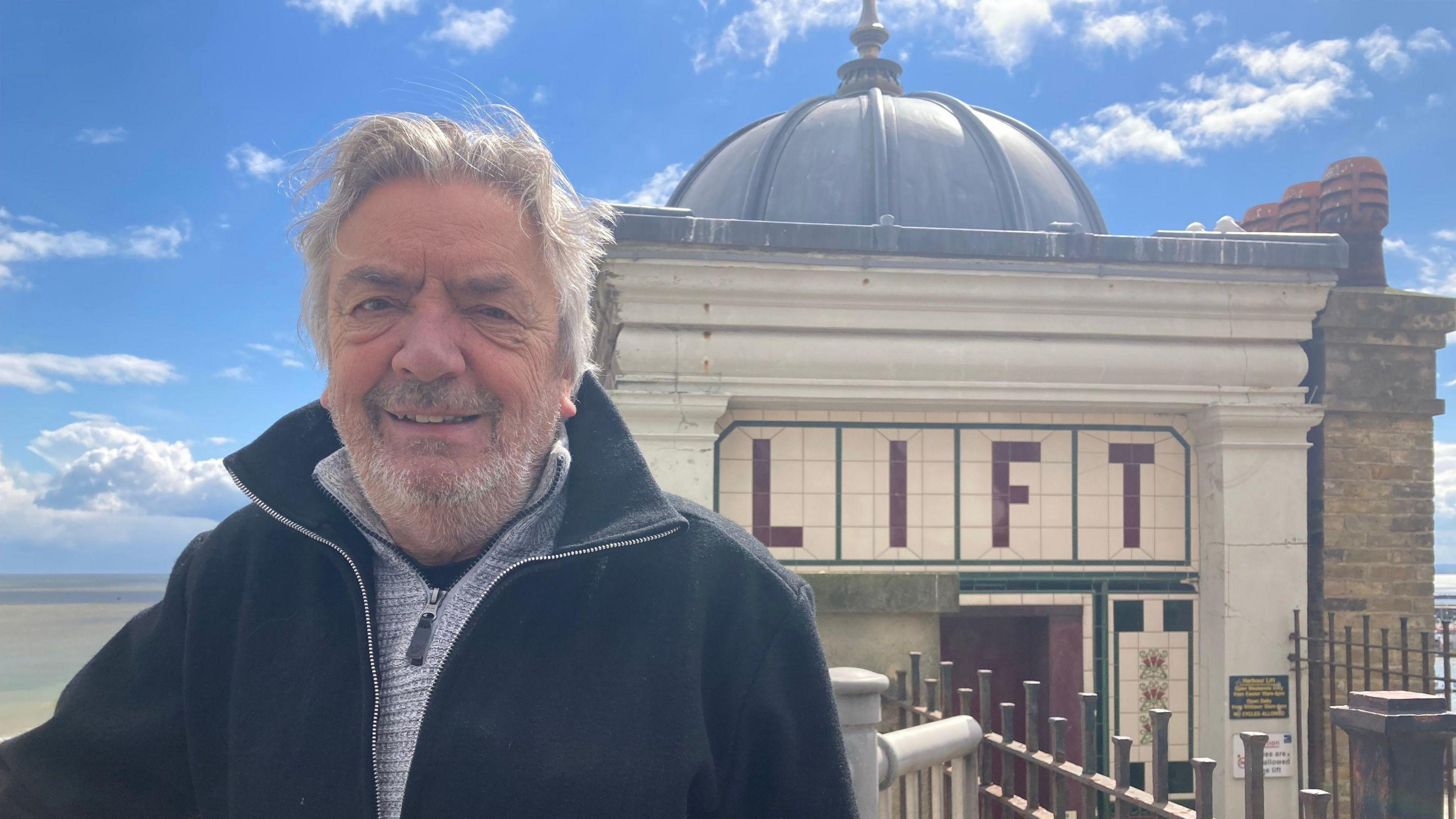 An older man with grey hair and stubble smiles at the camera in front of an historic lift in Ramsgate on a sunny day