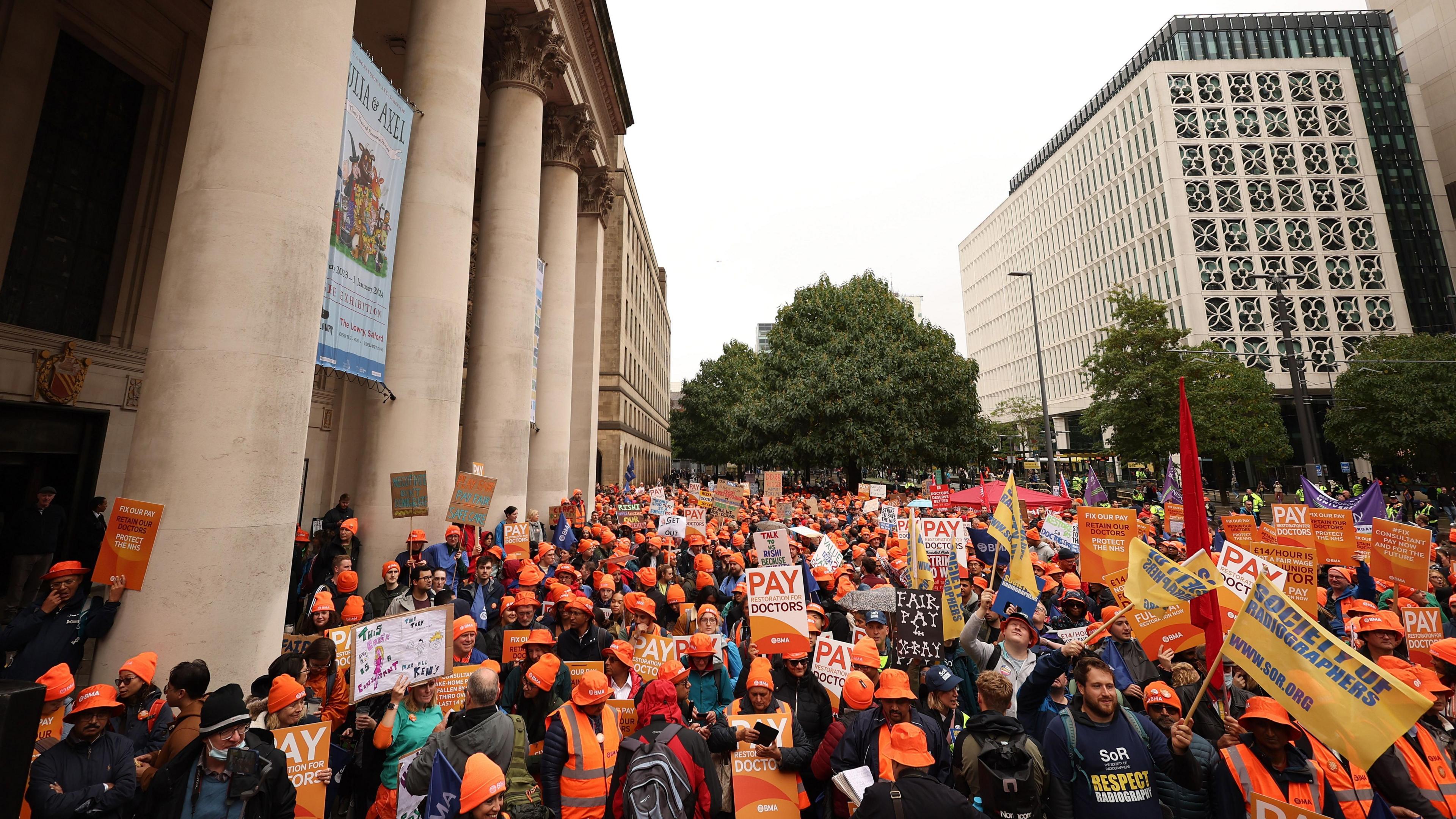 A large crowd of people in orange clothing and headgear waving flags and placards in support of the strike. They are next to the classical pillars at the library entrance. High-rise buildings and trees surround them at the public square.