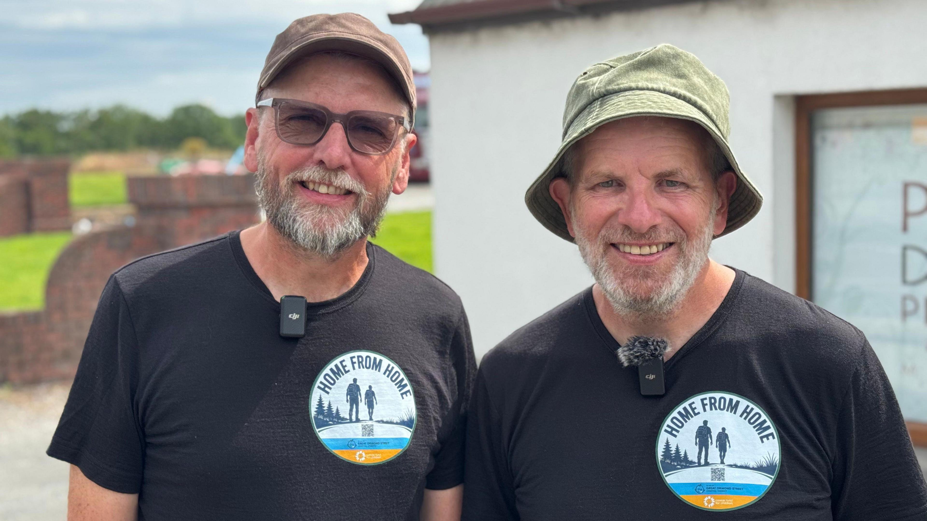 John and Eamon pictured together, both wearing black t-shirts and sporting short grey beards. One wears a green bucket hat and the other a brown peaked cap. 