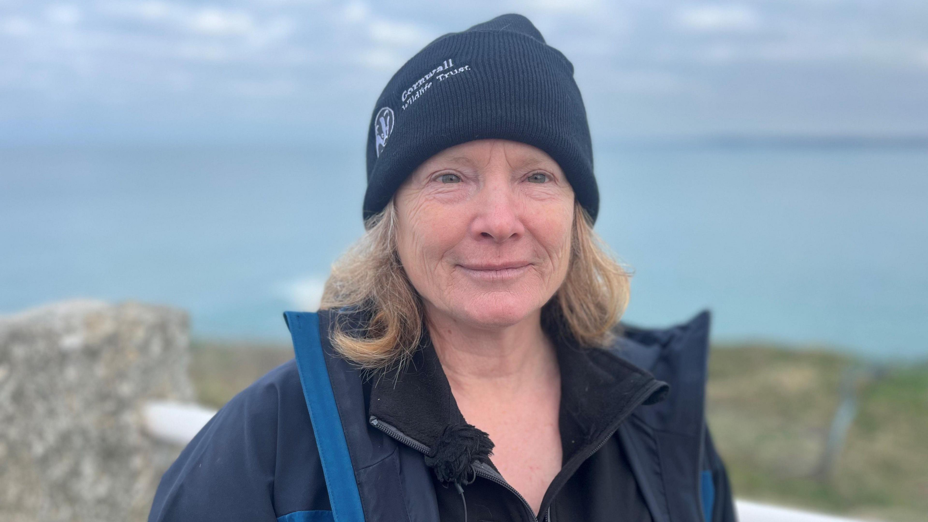 Bex Allen smiles at the camera. She is standing with Watergate Bay behind her on a clear day. She is wearing a woolly hat that reads CORNWALL WILDLIFE TRUST, a fleece and an anorak.