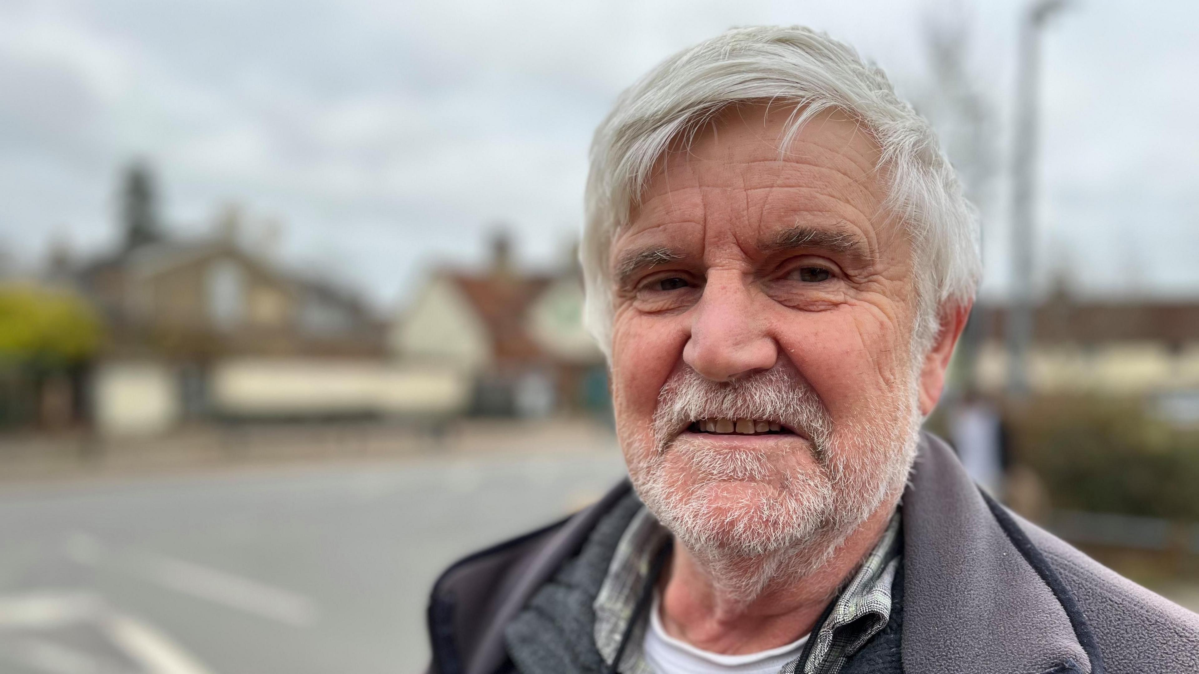 Brian Ross has short white hair and a short white beard.
He is wearing a grey fleeced jacket with a grey jumper underneath. He is smiling at the camera with buildings and a road behind him.