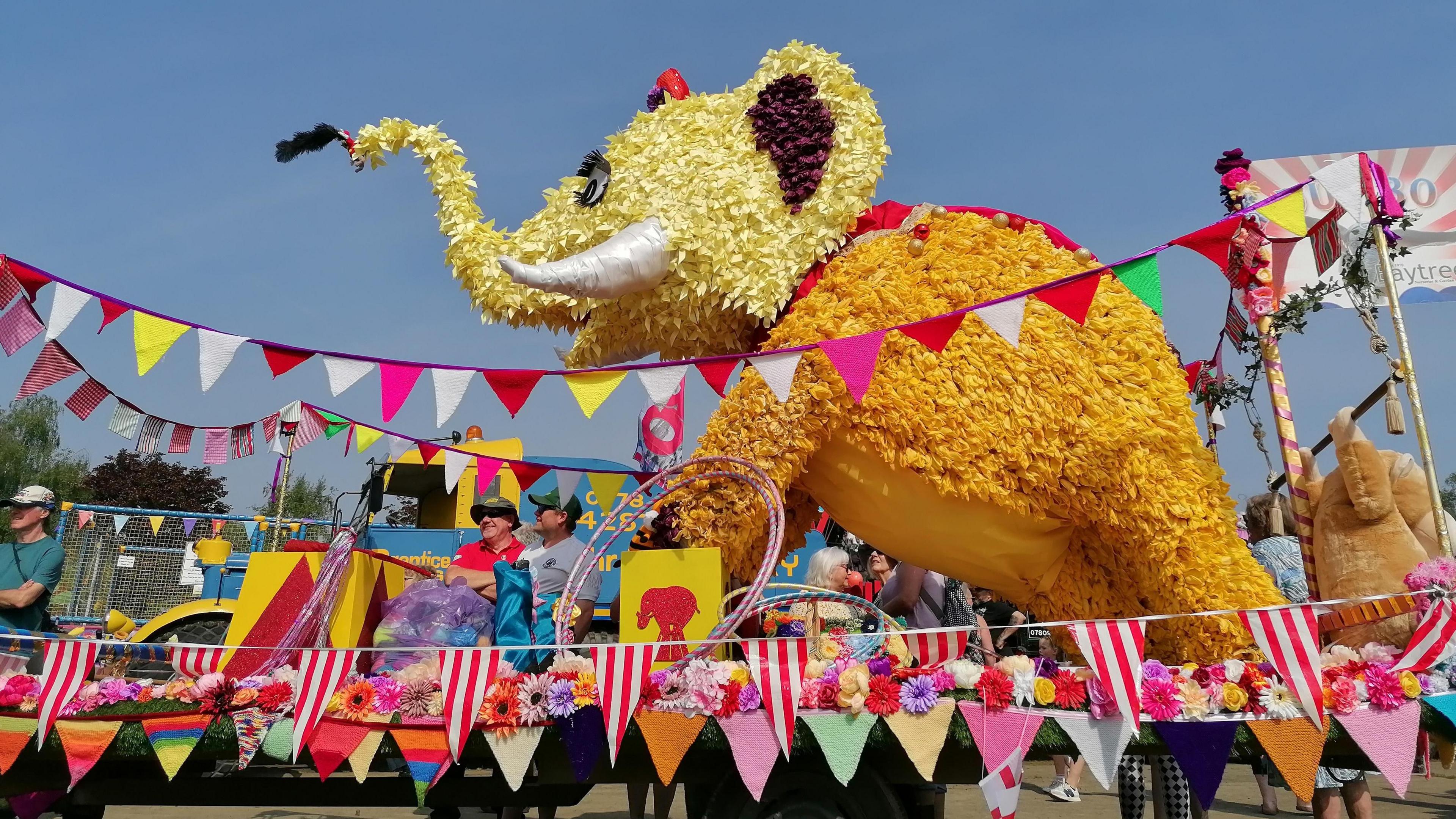 A large elephant on a float made from yellow tulip petals, it is surrounded by circus hoops, colourful bunting and more flowers. 