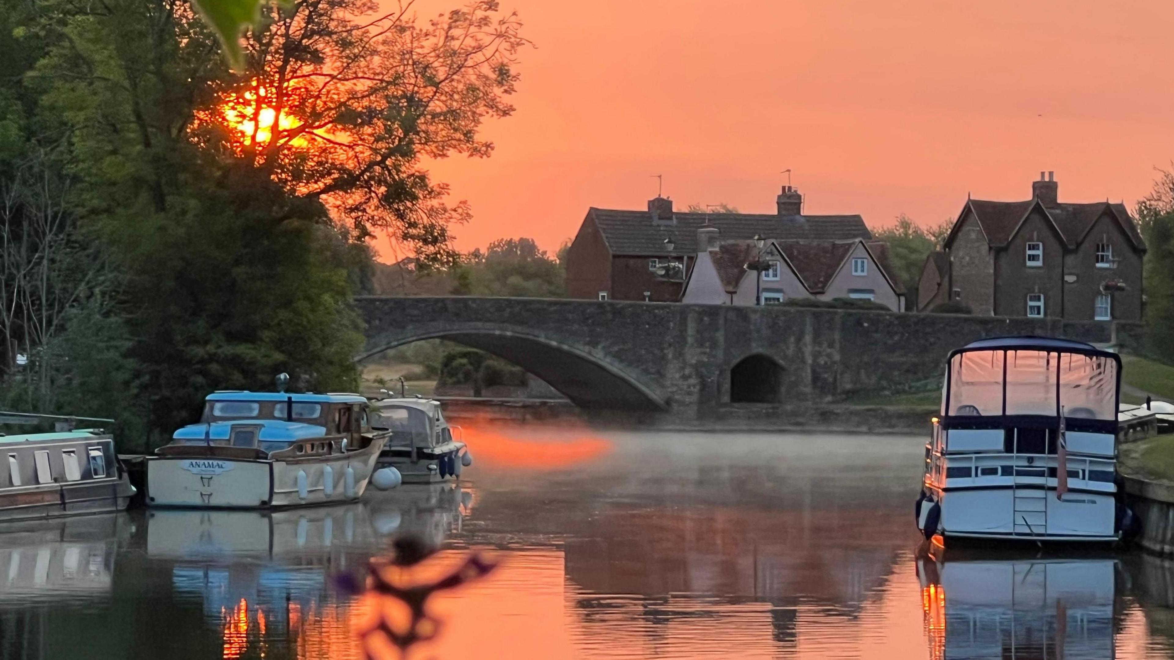 Small boats can be seen moored to river banks on either side of flat waters. A rising sun is providing weak orange glow across the sky and reflected in the water. An early morning mist can by seen on the surface of the water near to a stone bridge and some houses
