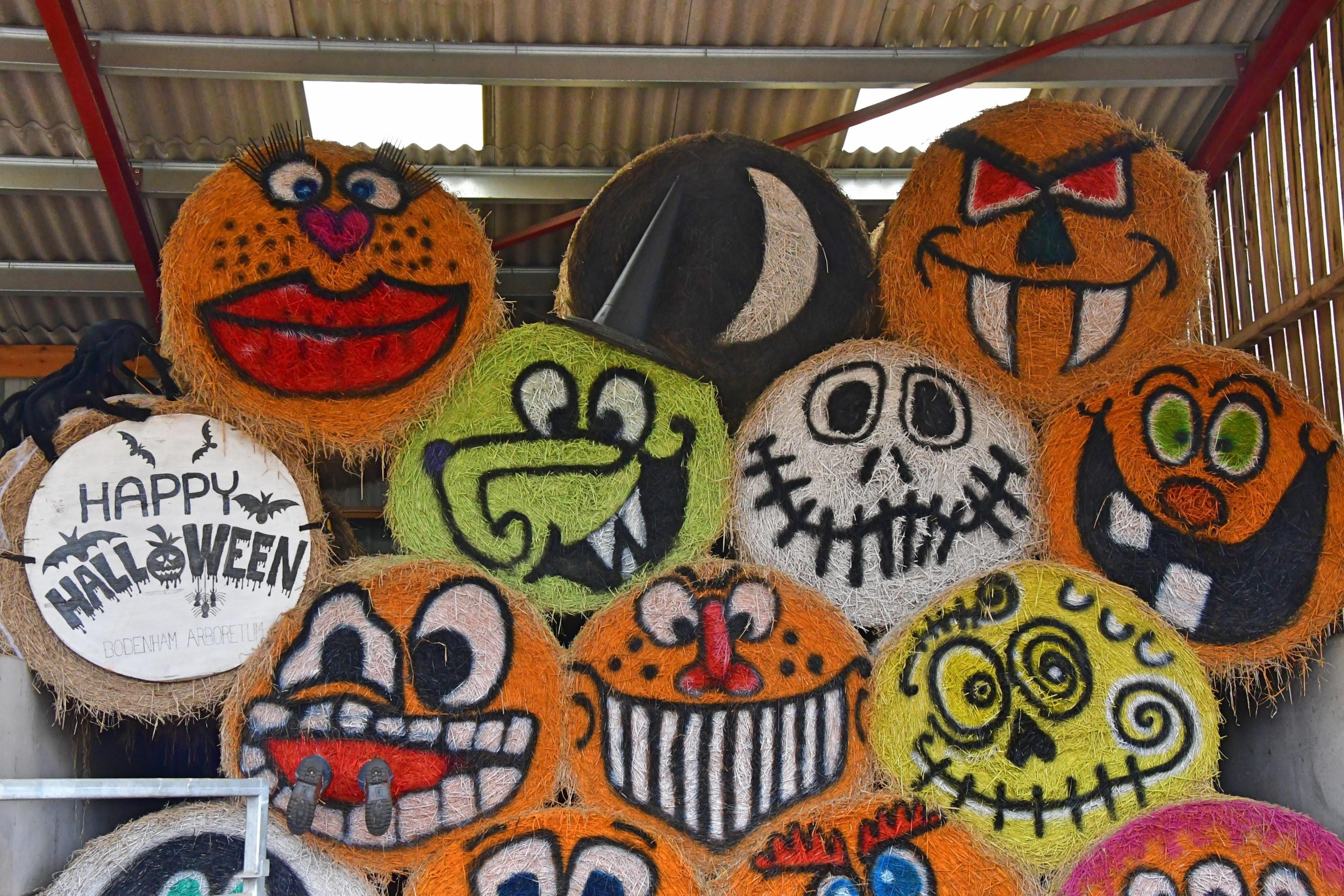 A stack of decorated around hay bales reaching up to a corrugated farm shed roof. They have faces mainly depicting carved orange pumpkins, but also skeletons and other Halloween themes. A sign on the furthest-left pumpkin reads "Happy Halloween".