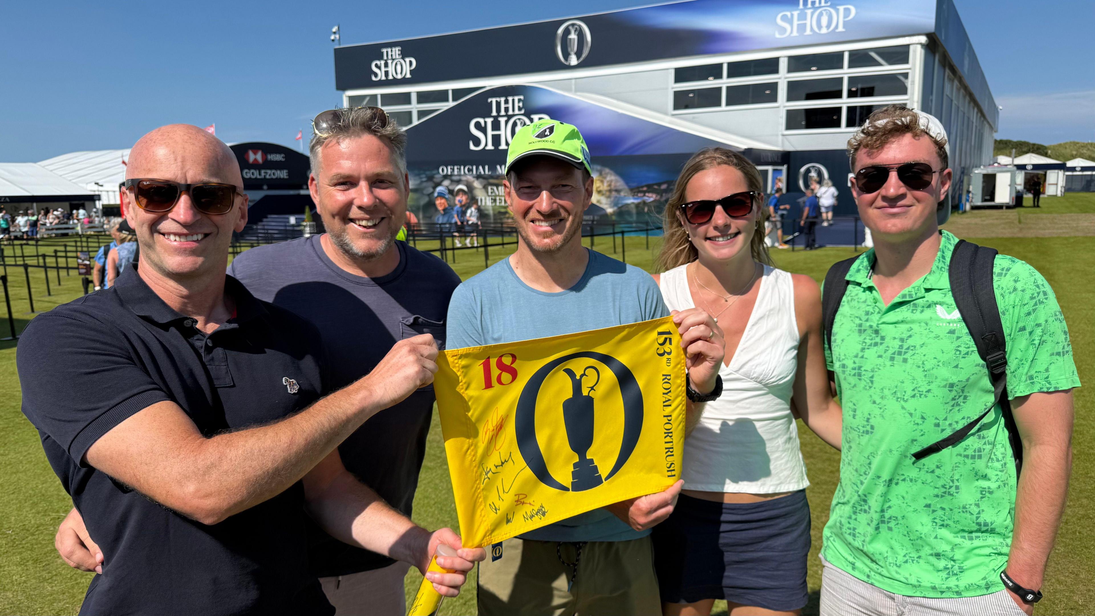 Five people standing in a line holding up the 18th flag. It is yellow with The Open branding in navy.