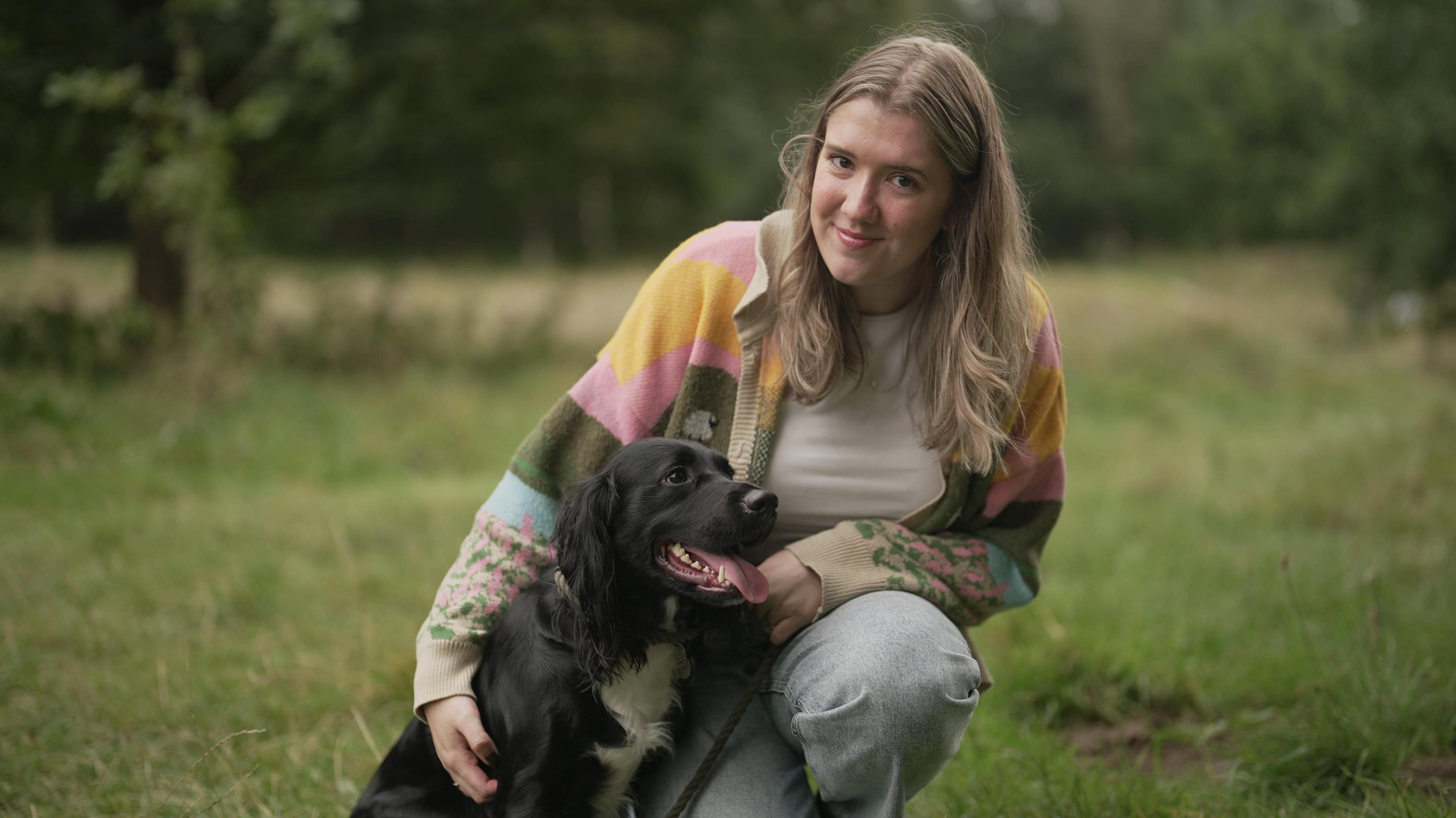 A woman with blonde hair wearining a patterned cardigan and jeans is crouched down on the grass next to a black dog. The dog has its tongue sticking out and has a white patch of fur on its neck.