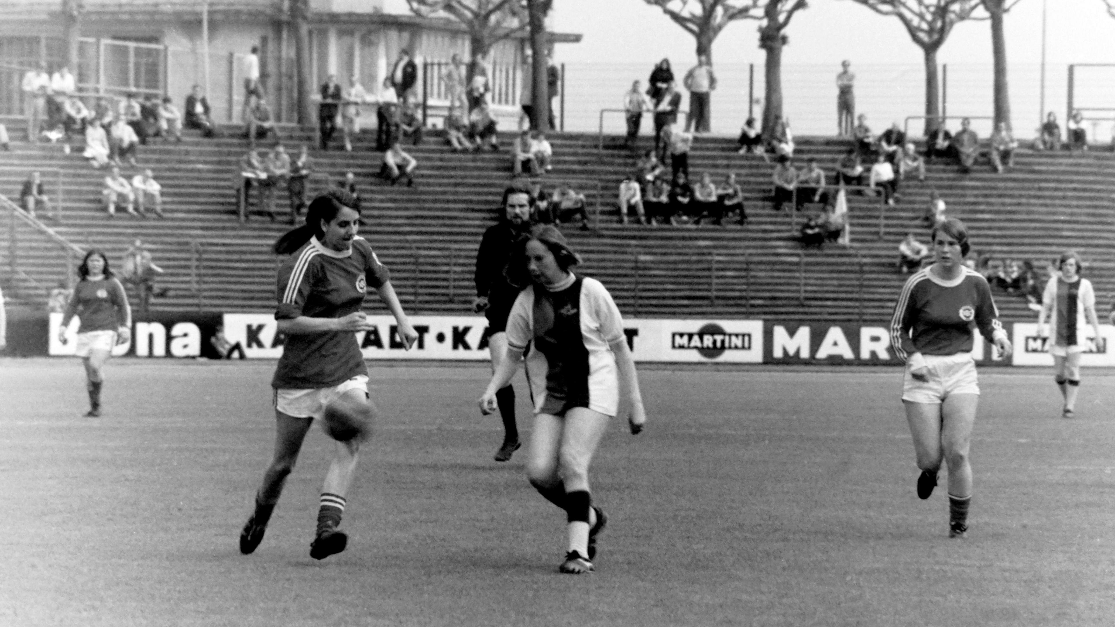 A black and white photo of a women's football match in the 1970s, featuring players from Crystal Palace Ladies in action on the field. Three players are in focus, with one player attempting to kick the ball while others look on. The background shows a nearly empty stadium with a few spectators seated on the stands.