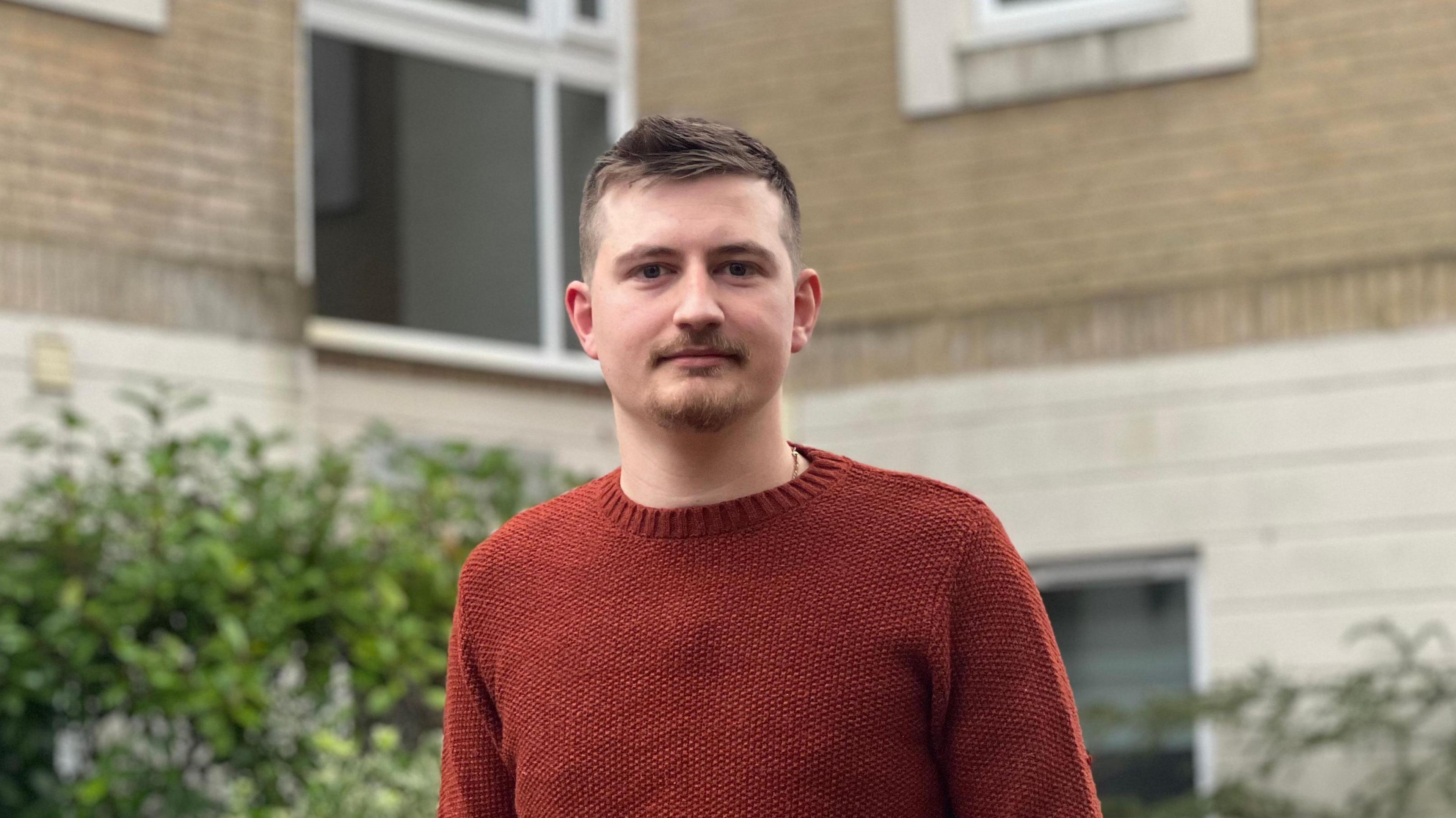 Matthew, wearing a bronze-red jumper, stands in the foreground, with blurry plants and his block of flats in the background 