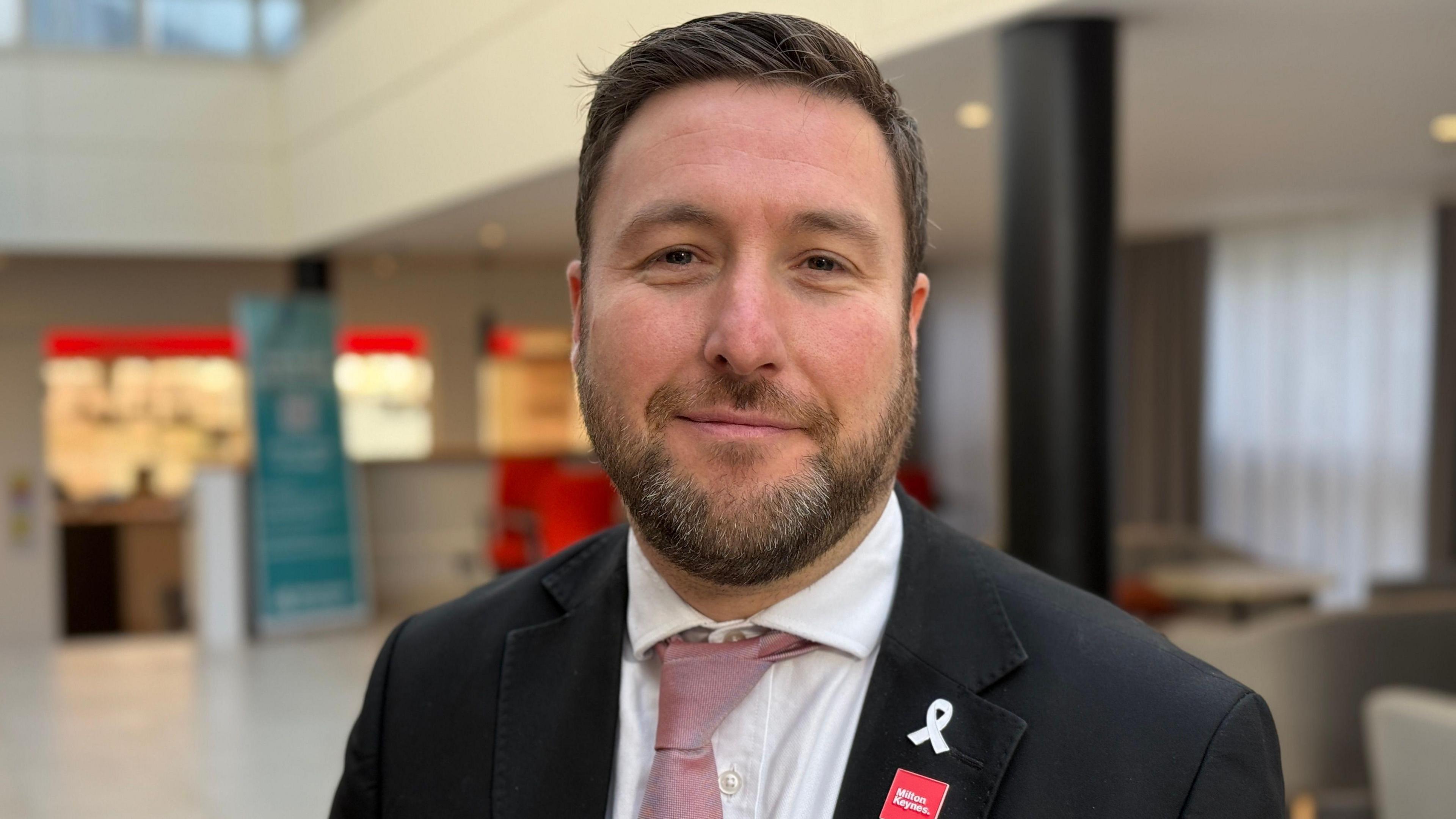 A head and shoulders shot of Pete Marland looking at the camera and smiling. He is wearing a suit jacket, a white shirt and light red tie. A hotel lobby is blurred in the background.