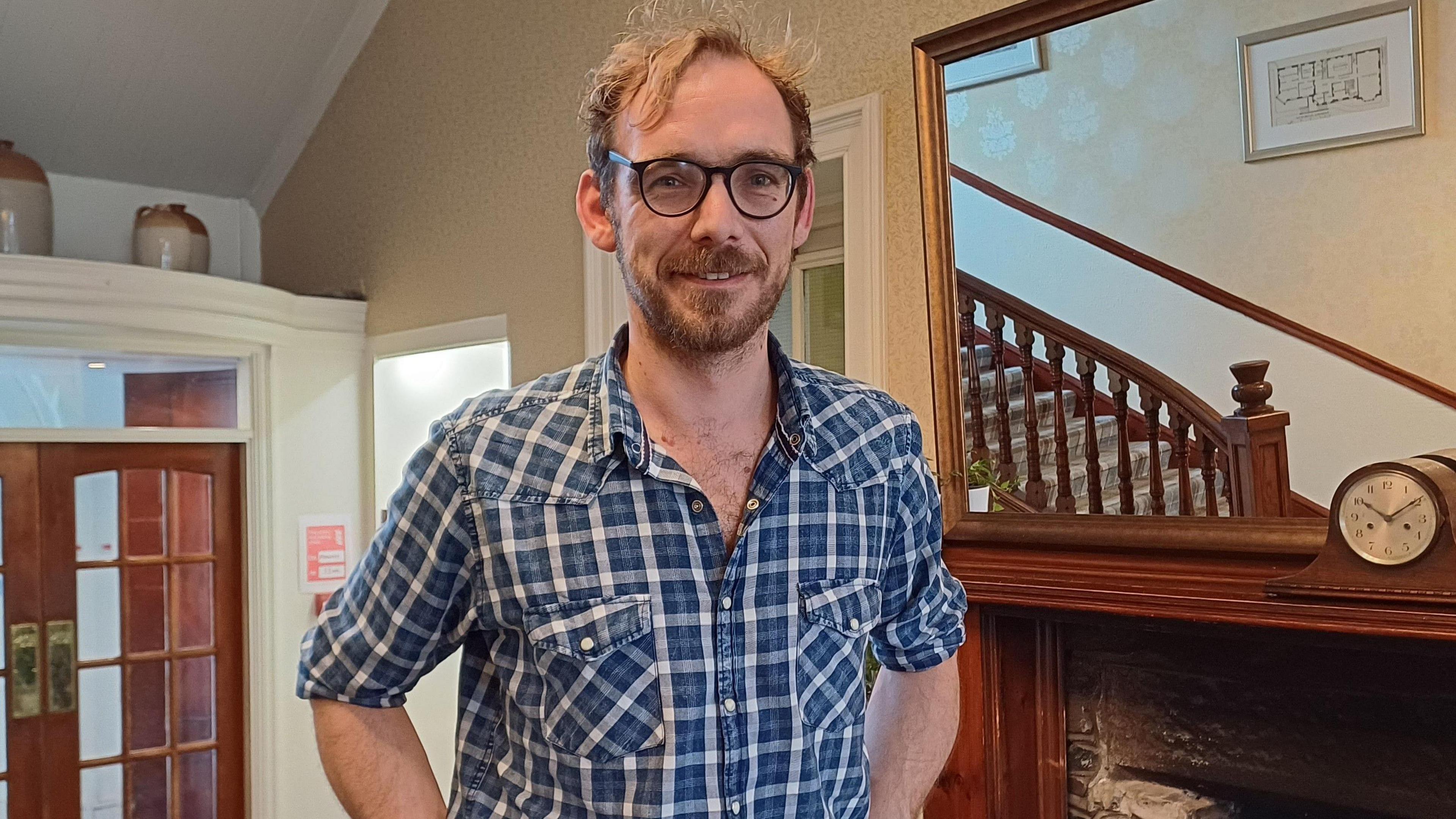 Bryan Botha is standing in the reception of the Western Isles Hotel. He has fair hair and a beard and is wearing glasses. He is wearing a blue and white checked shirt with the sleeves rolled up and is smiling.