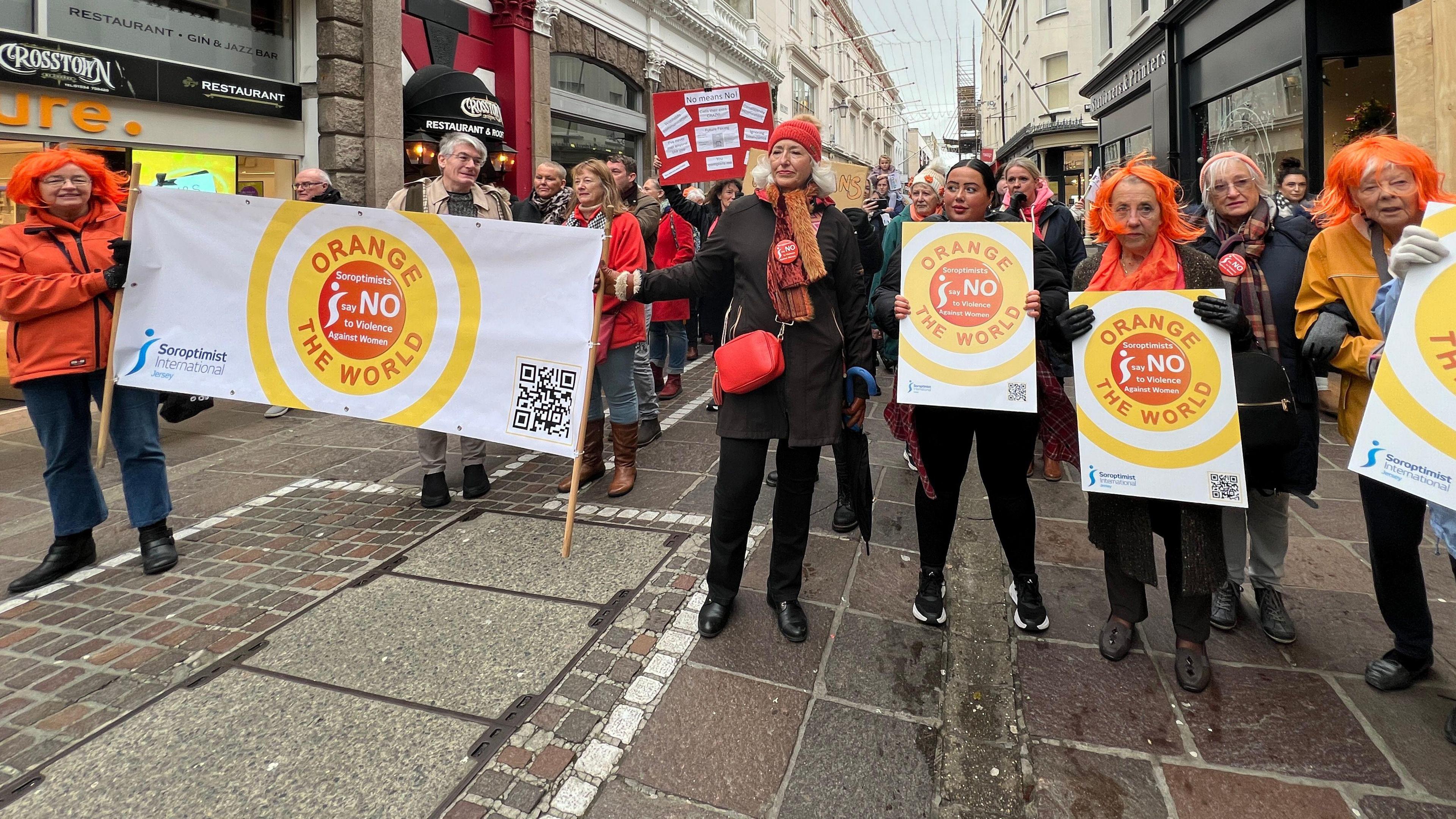 A group of women hold signs saying no and orange is the world. Some are wearing orange wigs as well. Many are wrapped in scarfs. They're standing on the high street in St Helier in Jersey.