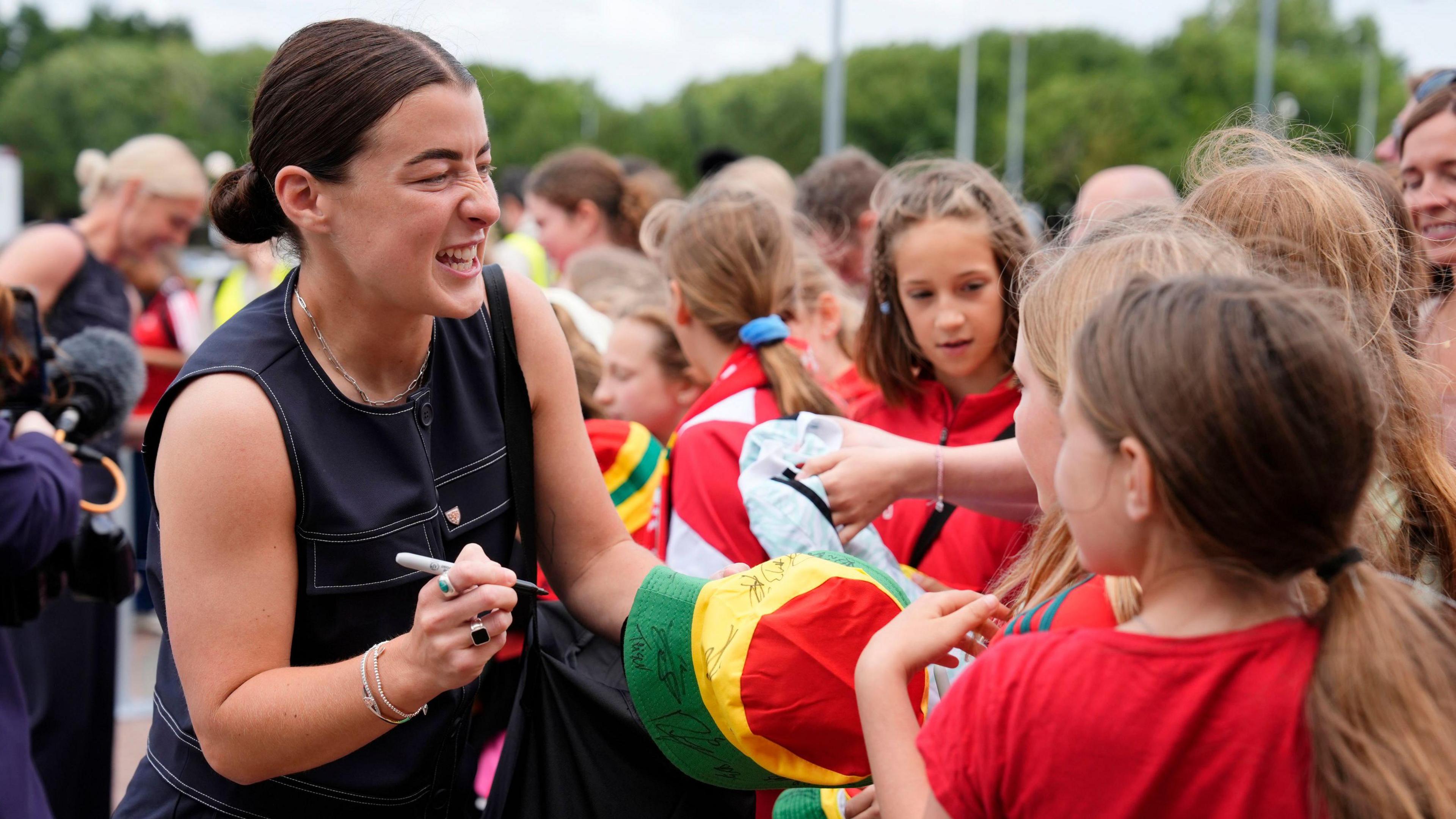 Angharad James (left) wears a black sleeveless top while signing autographs on a red, yellow and green hat for young fans on the right.