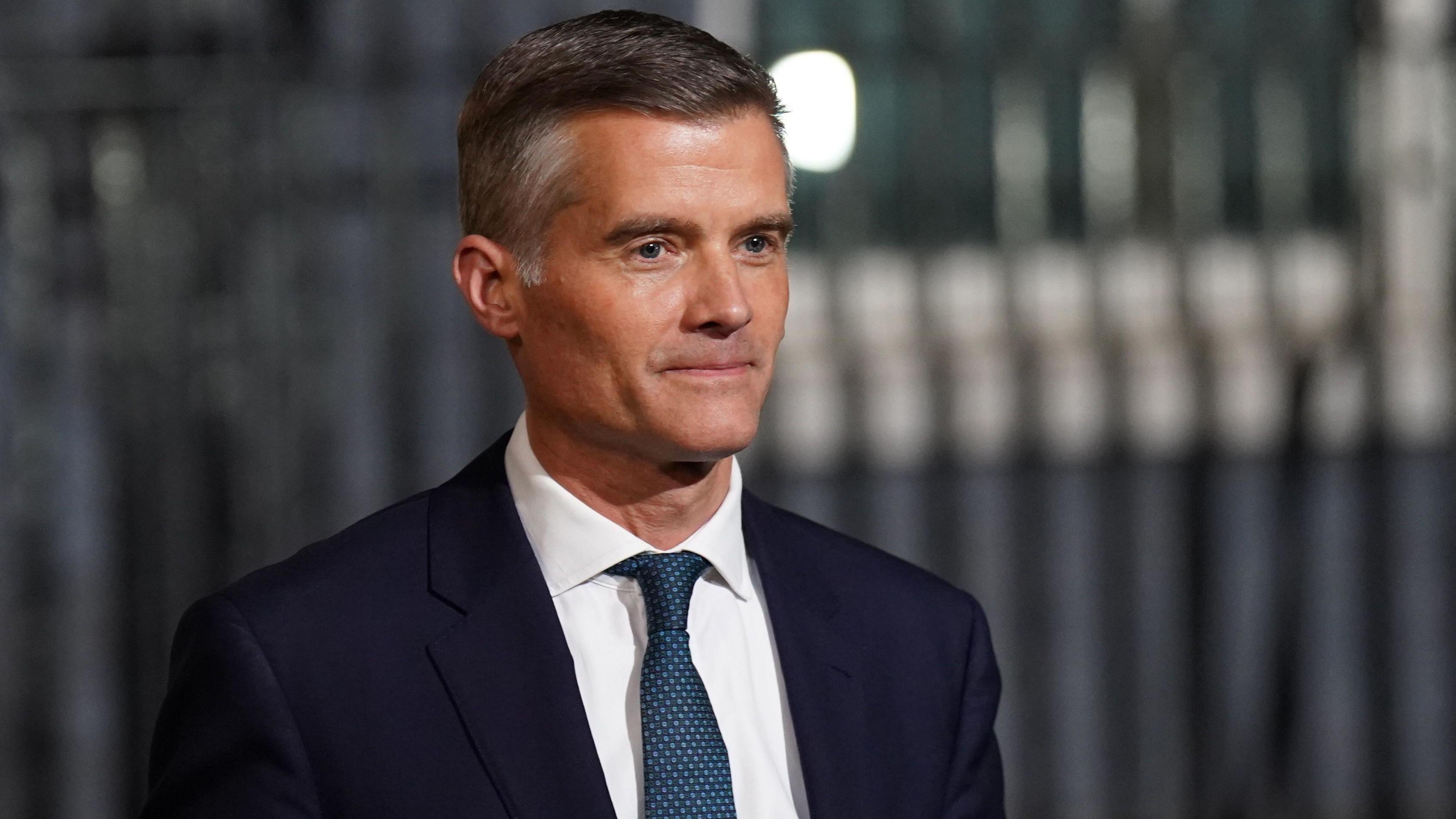Image of Mark Harper outside of number 10. The background is blurred. Harper is wearing a dark suit and blue tie.