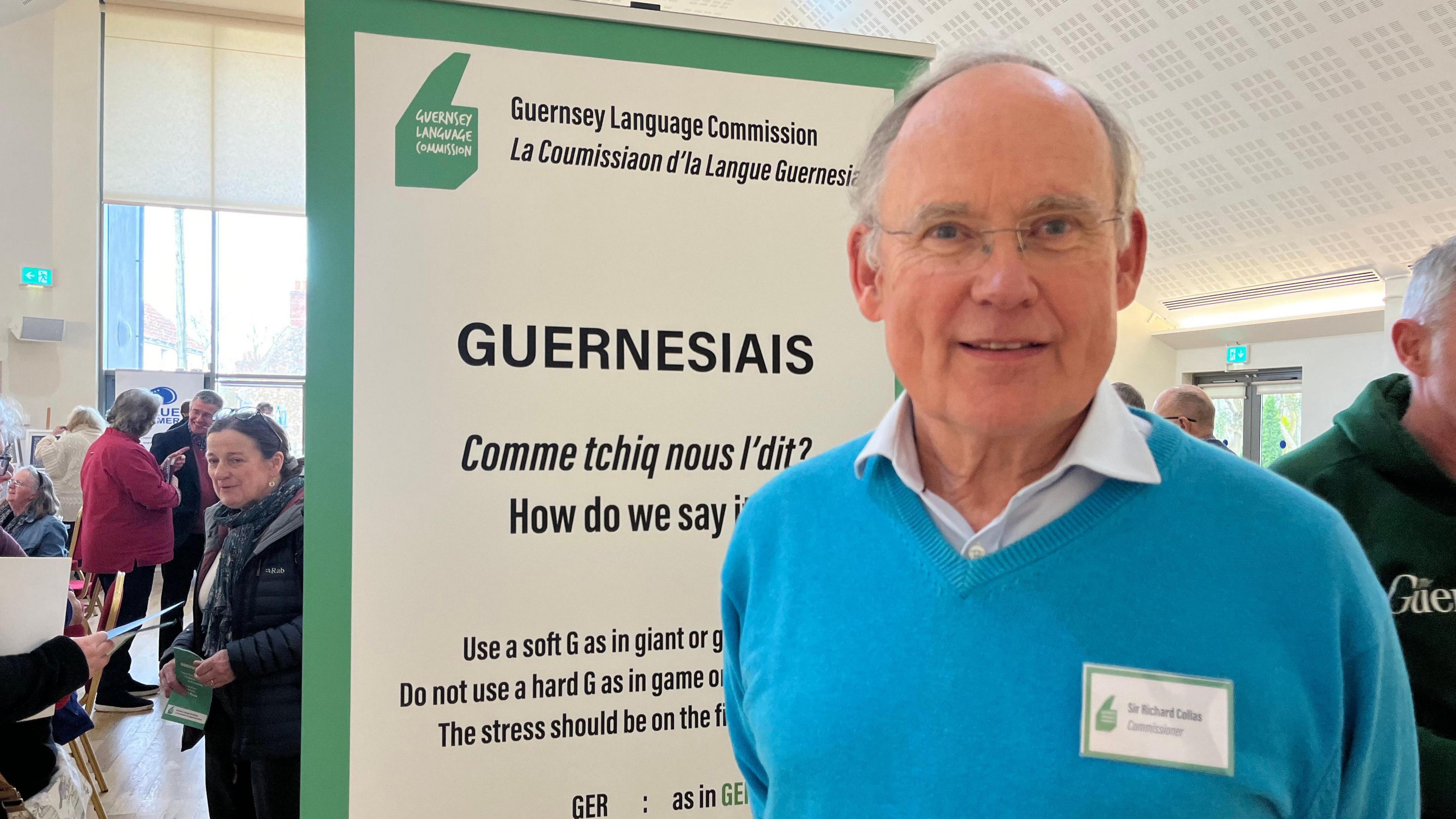  Sir Richard Collas the chair of Guernsey Language Commission stands in front of a banner explaining how to pronounce the word Guernesiais, using a soft g as in giant.