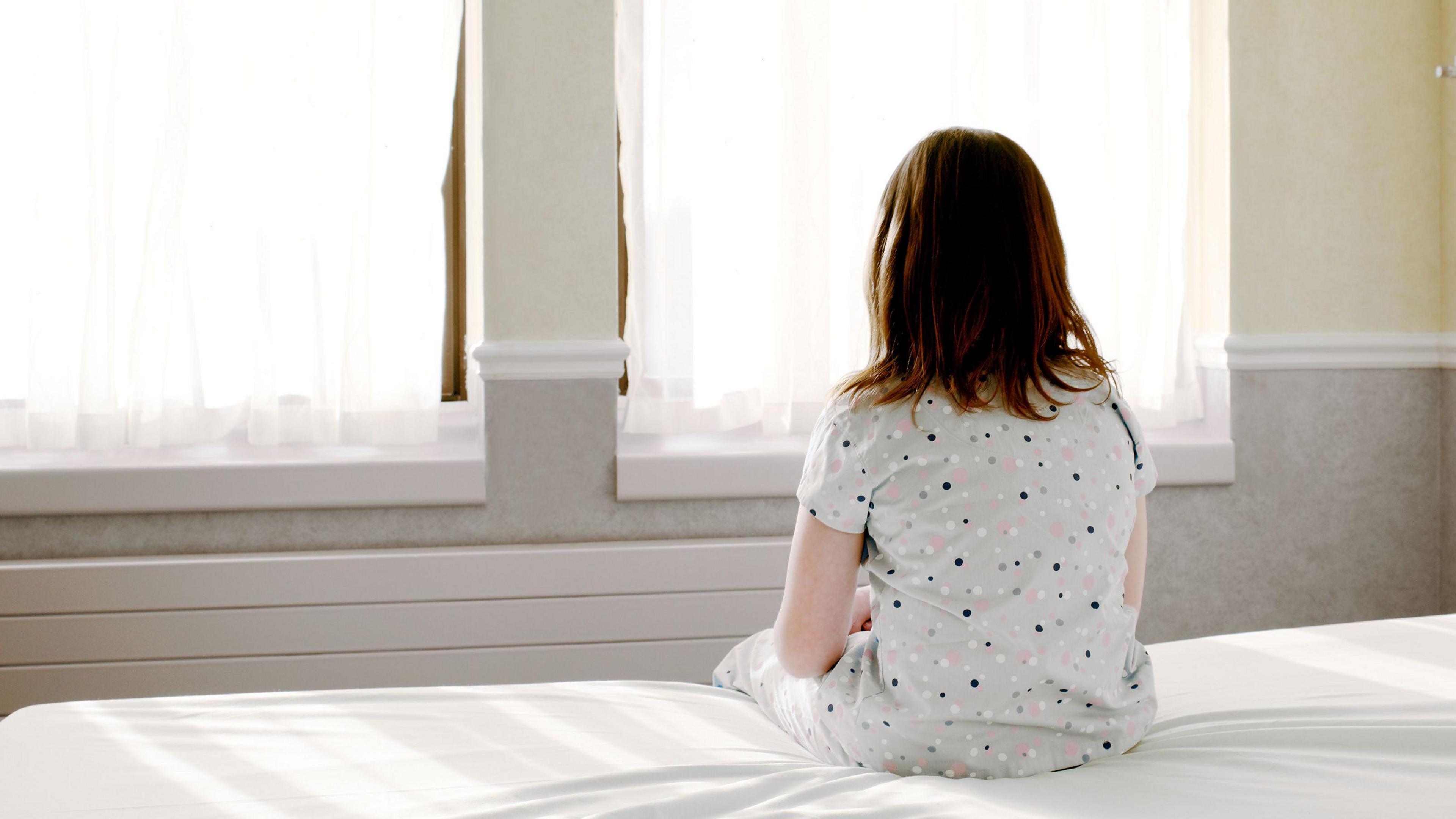 Back view of a young child sitting on a bed and looking towards a window