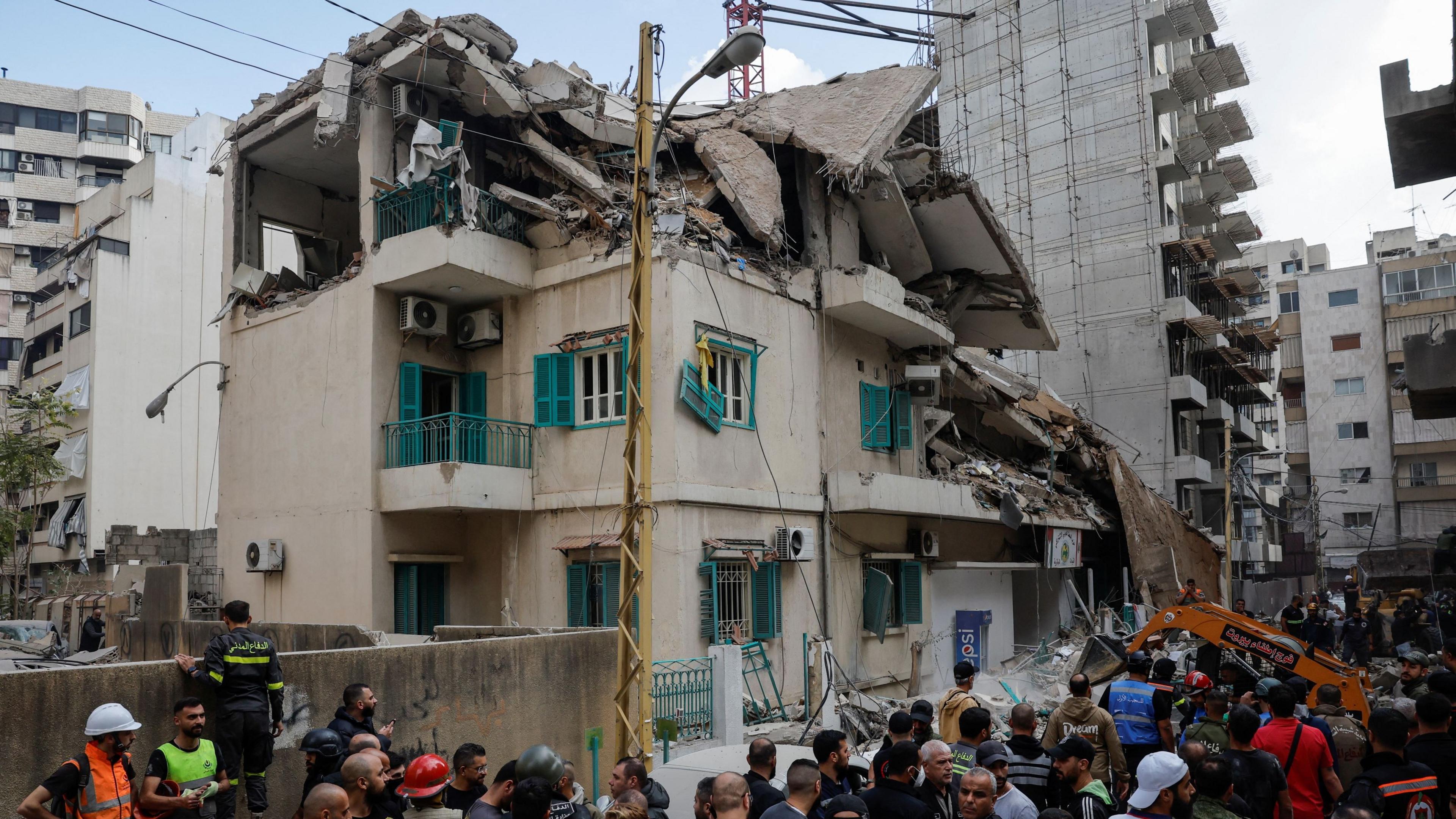 The roof of a multi-story building with tan walls and green balcony railings and window shutters is collapsed from an air strike, with a crane and a crowd of rescuers and onlookers below 