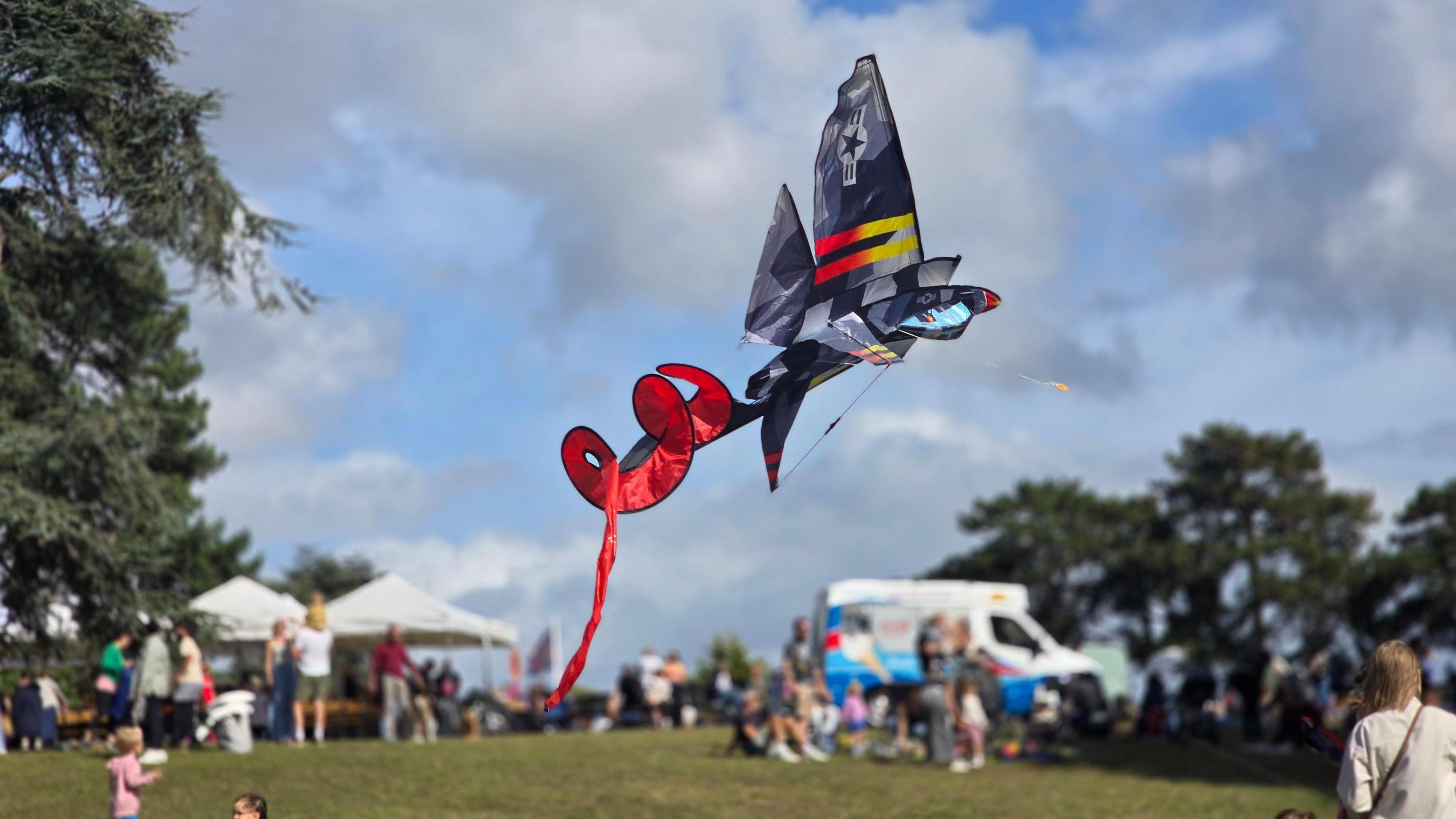 A dark grey kite shaped like a plane with a red long tail spiralling behind it flying in the sky. It is close to the ground as people can be seen on the grass in the background along with an ice cream van, some gazebos and trees. The sun is shining with lots of clouds in the blue sky.