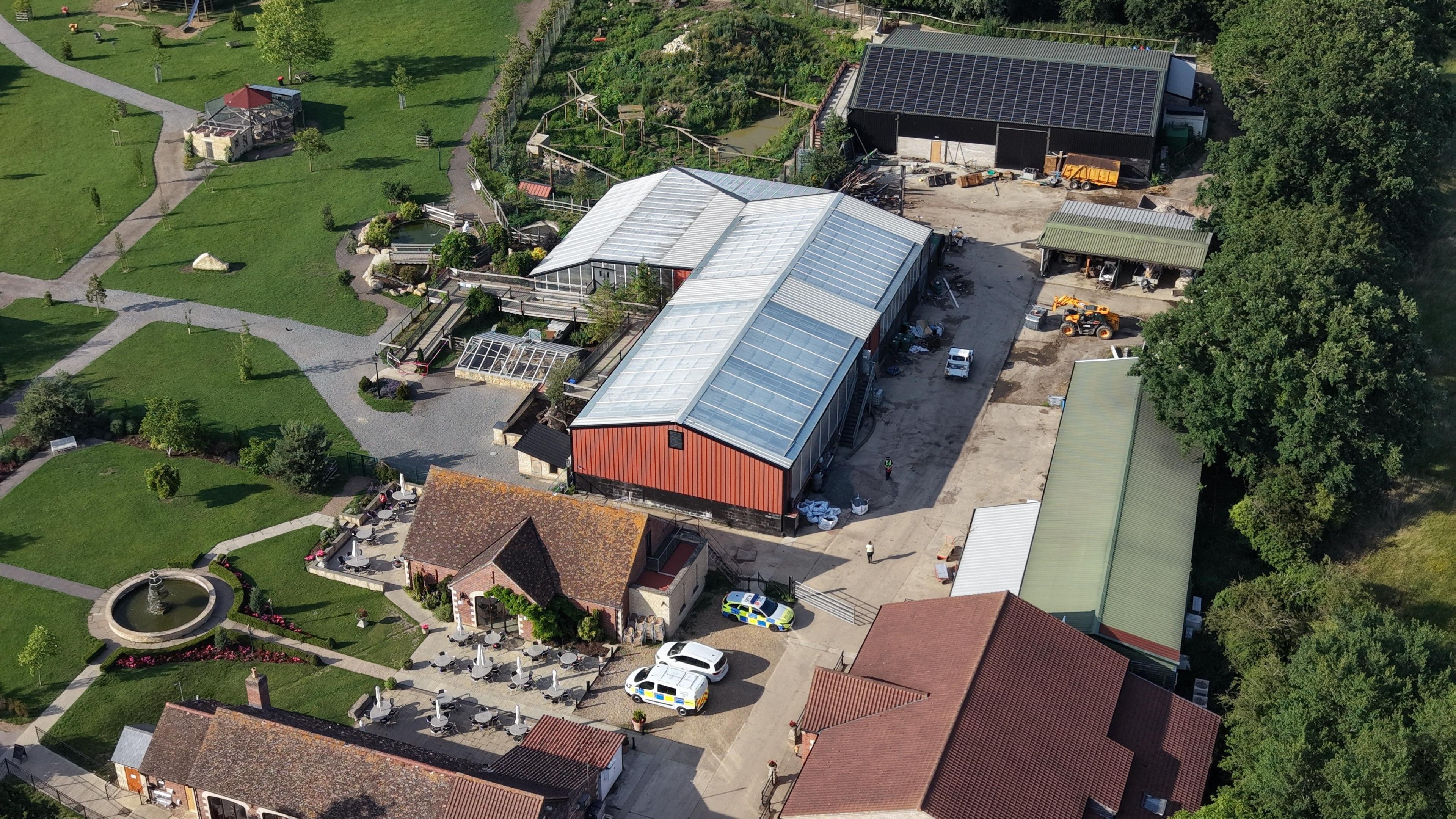 A drone image from above a farm and zoo. There are several large buildings housing enclosures as well as outdoor areas. Some vehicles can be seen parked around the site.