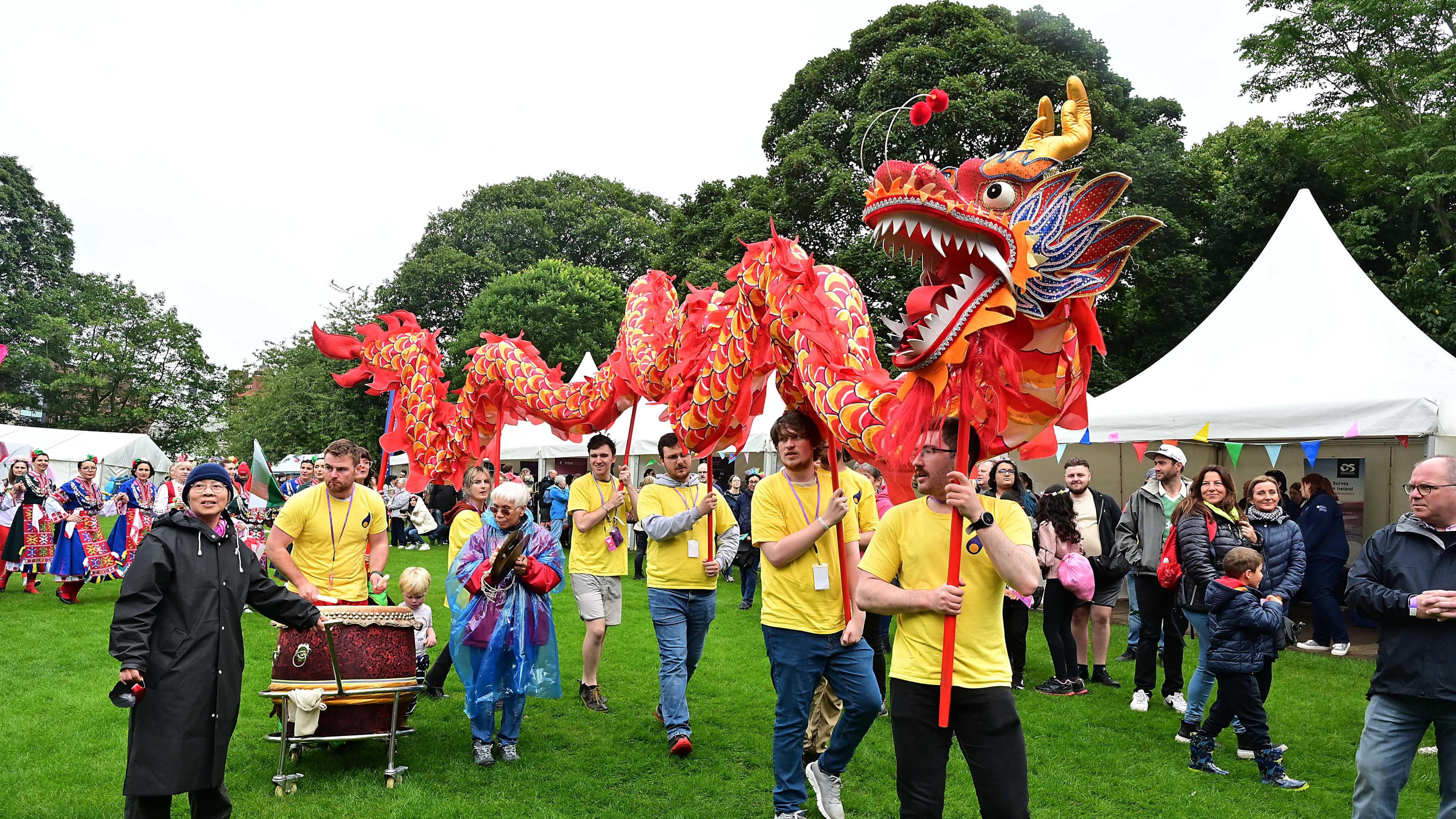 A dragon puppet being carried through Botanic Gardens for Mela Day. It's a large red, gold, blue and orange puppet which has it's mouth open showing white teeth. It is beinbg carried by six people wearing yellow tshirts and holding the puppet above their heads on sticks
White tents and attendees appear in the background