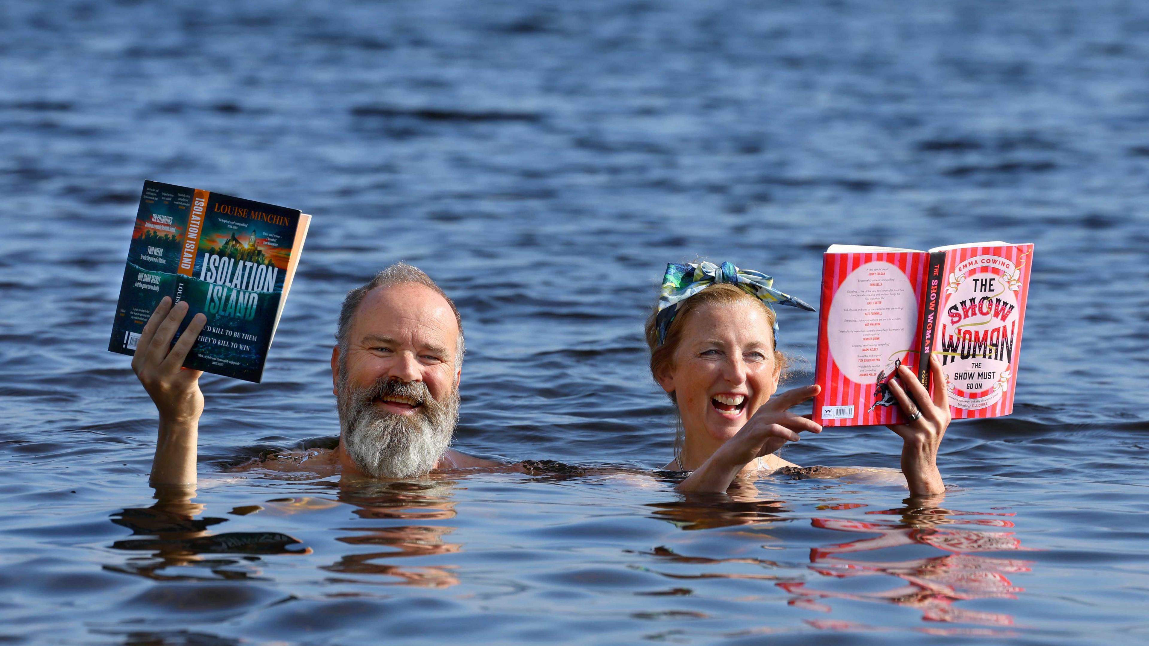 Greg Hemphill and Julie Wilson Nimmo in water. They are both holding open books in the hands and smiling at the camera.