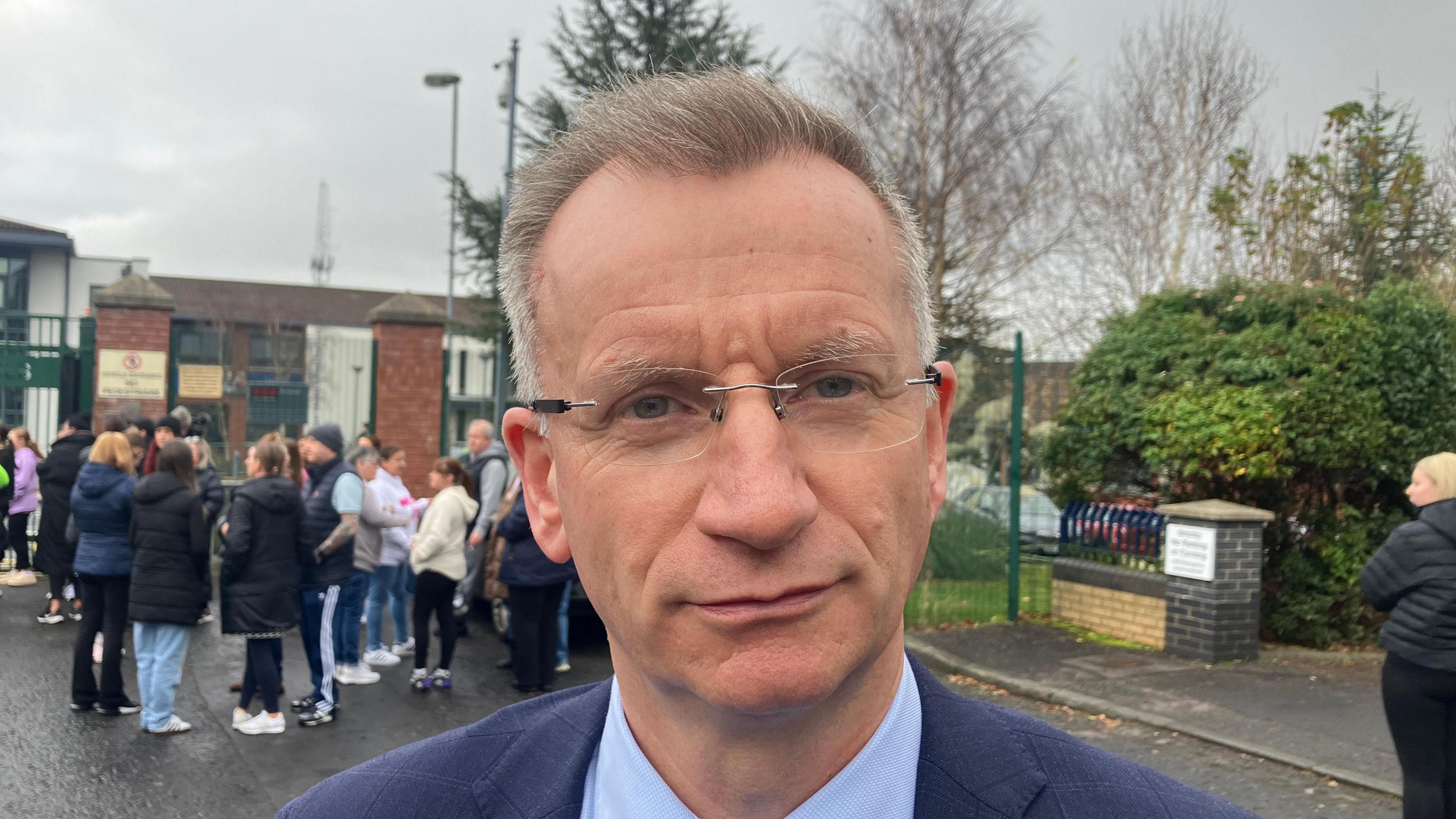 A close-up shot of Kingston. He has short grey hair and is wearing glasses and a navy blue suit.