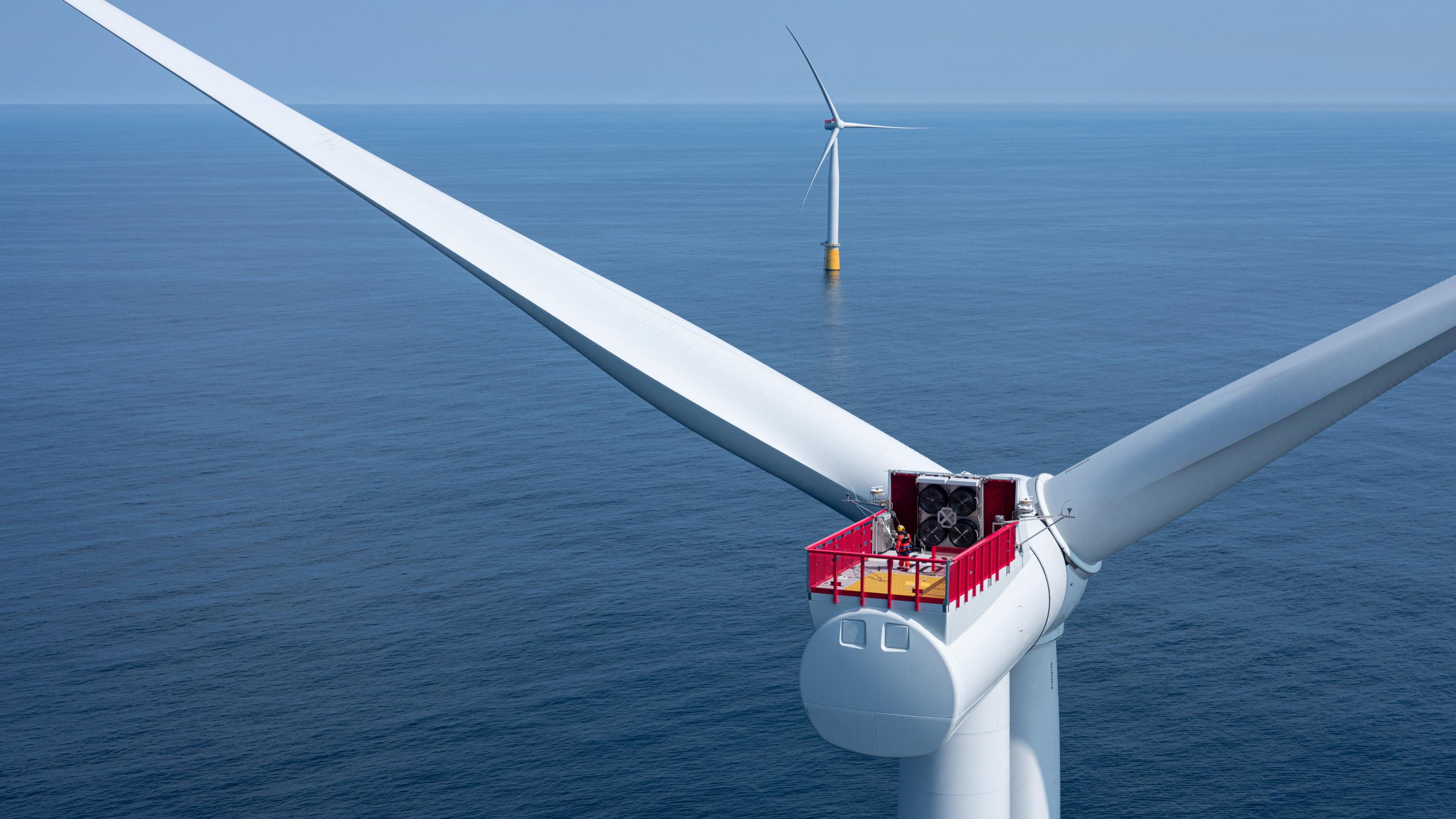 The rotor of a large wind turbine at sea. The turbine is white and there is a red and yellow platform over the axel with two people in hard hats wearing red on it. In the distance another wind turbine stands in line with it.